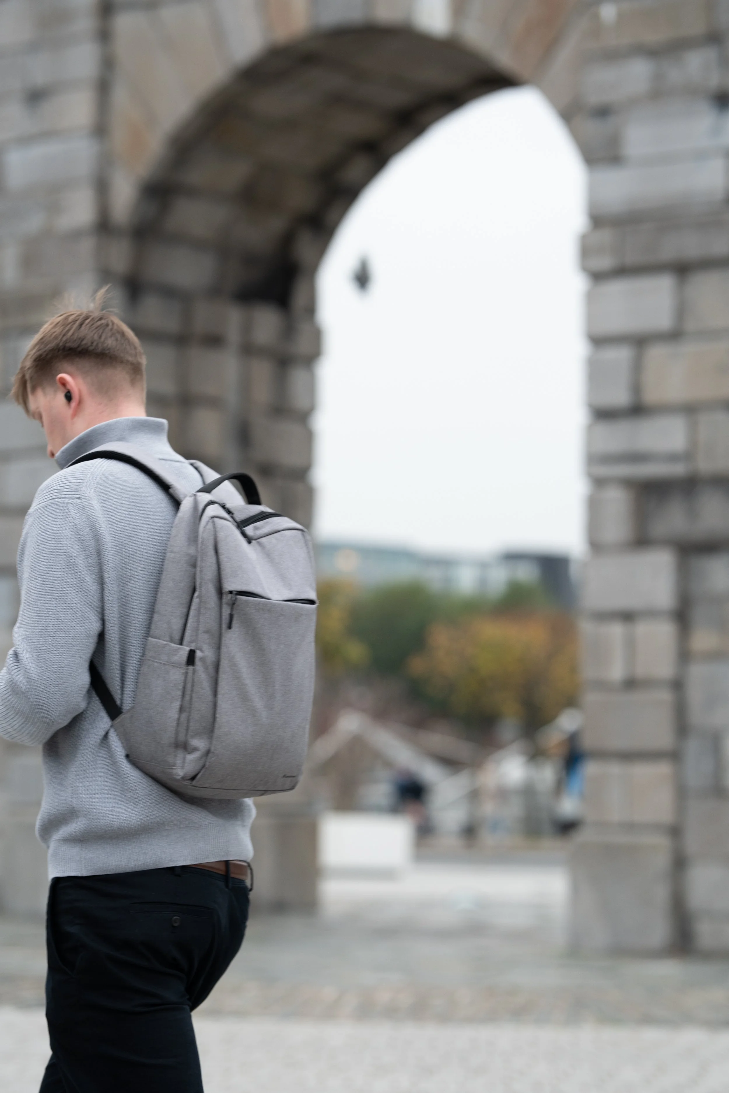 Young man in gray sweater with a gray backpack standing near stone archway, looking at his phone.