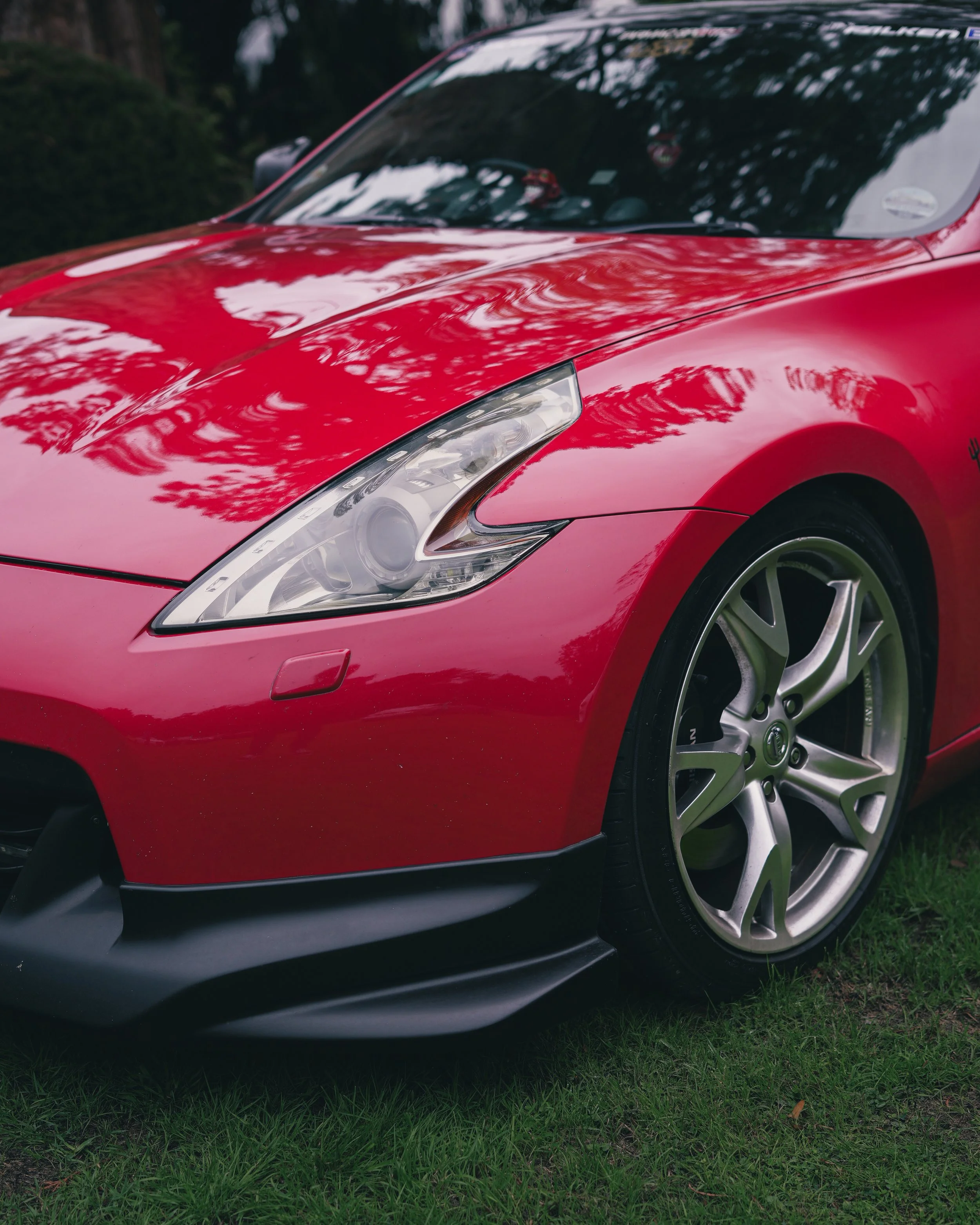 Close-up of a red  Nissan z370 sports car's front end, showing headlight, front tire, and black lower bumper with reflections of trees on the hood.