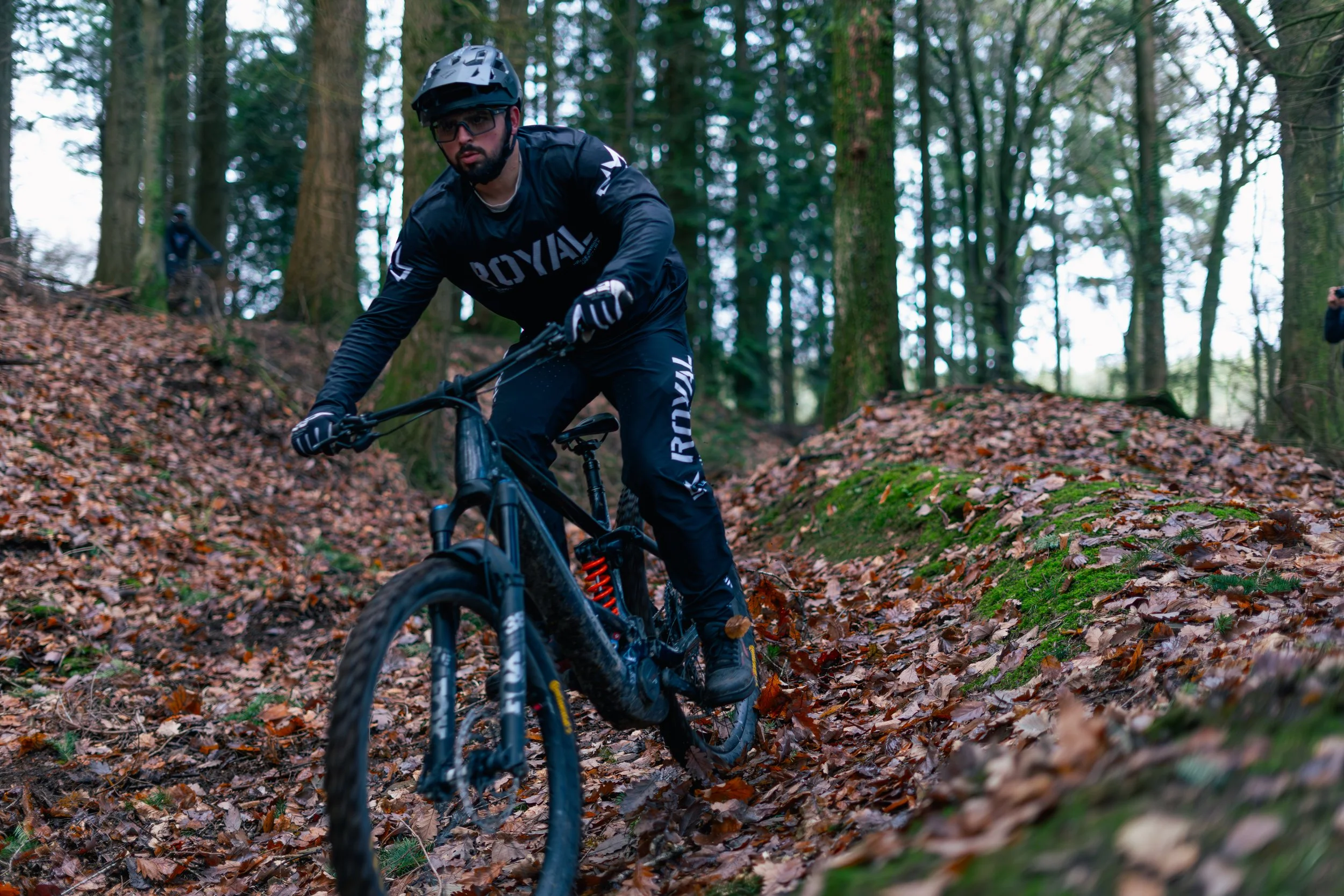 A man mountain biking on an outdoor trail through a forest, wearing a helmet, sunglasses, black riding gear, and gloves, with another cyclist visible in the background.