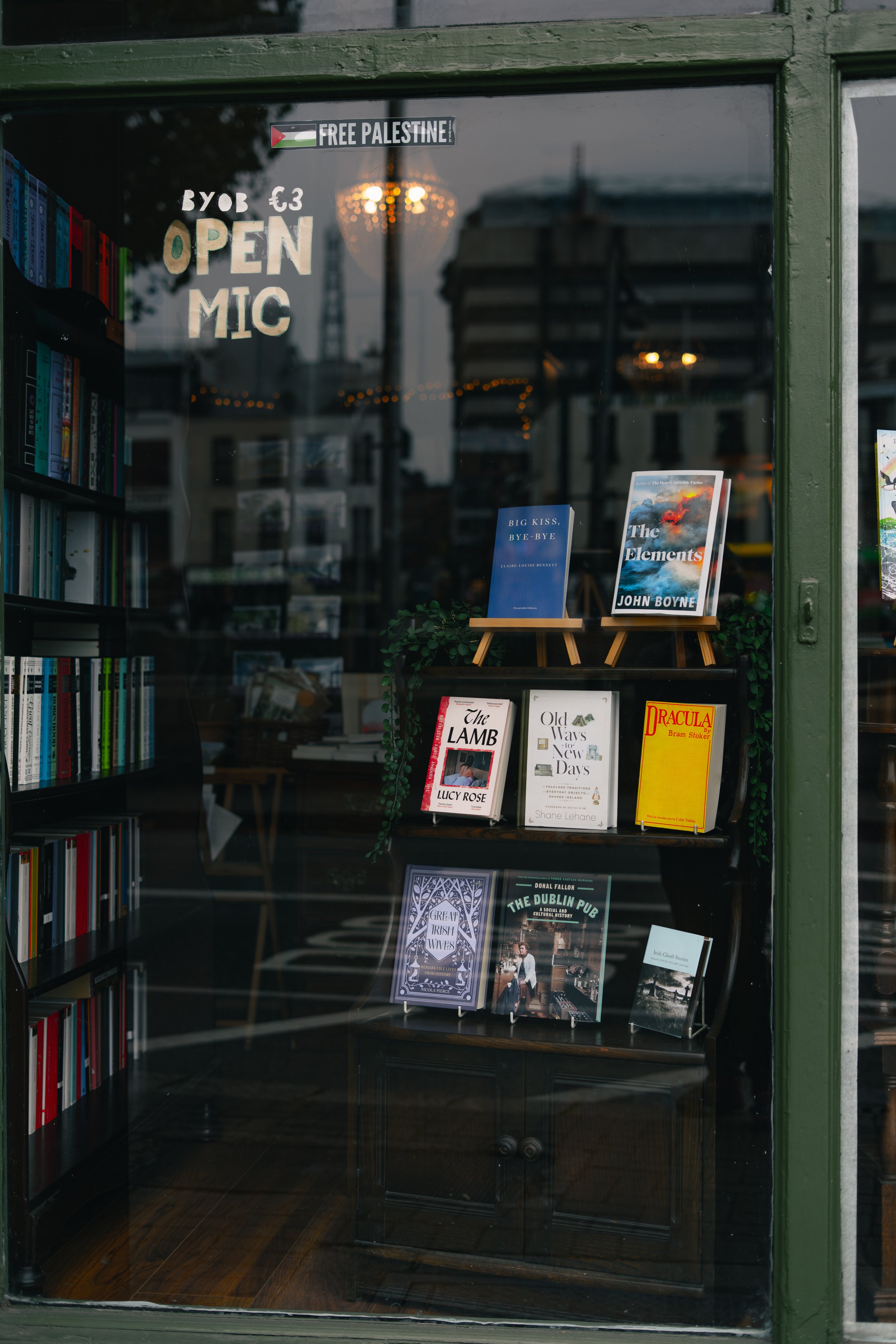 View of a bookstore window displaying books on shelves and stands, with signs indicating an open mic night for 3 euros, and a sticker reading 'FREE PALESTINE'. The window reflection shows buildings and a street outside.
