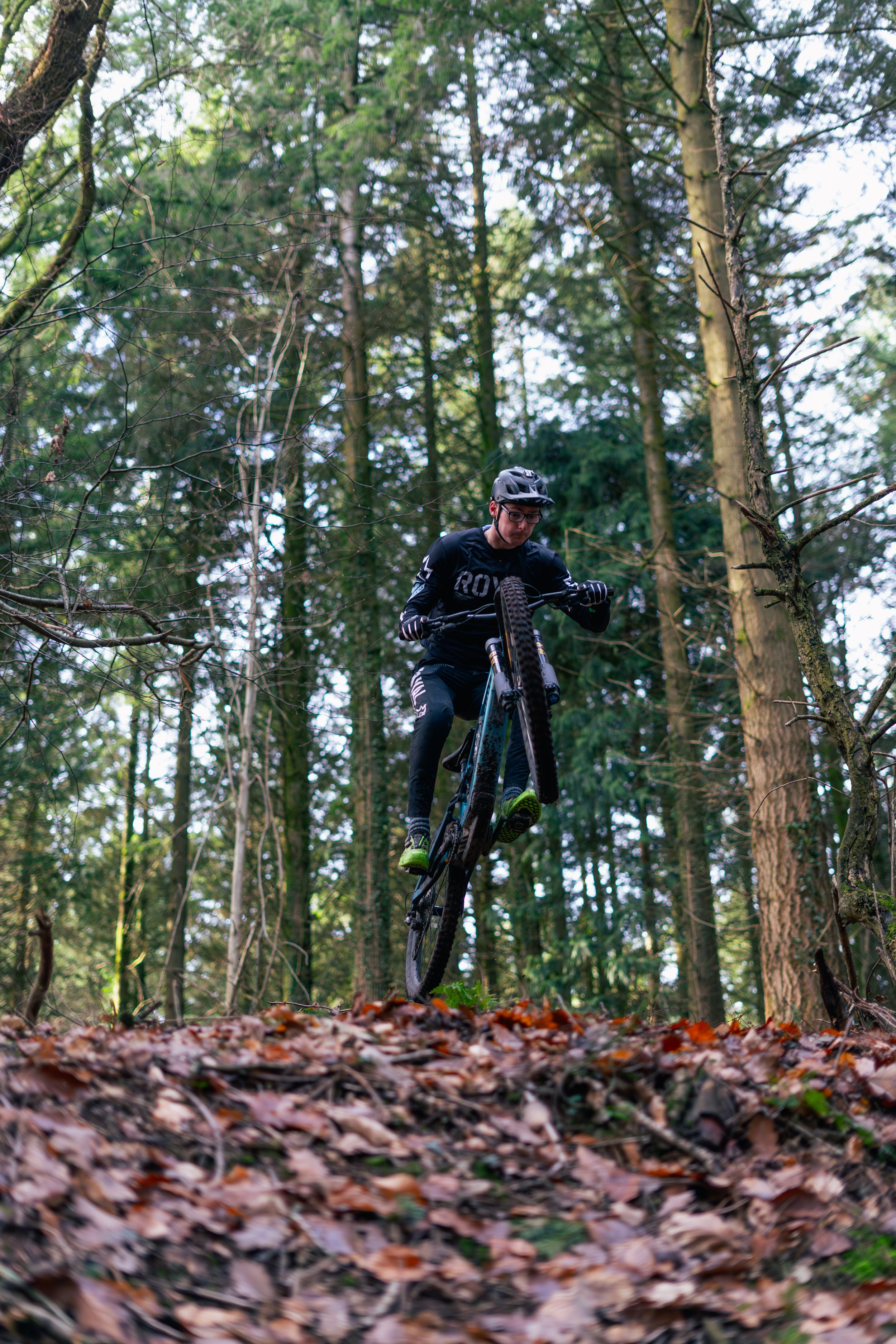 A person in black riding gear and helmet performing a jump on a mountain bike over a leaf-covered forest trail.