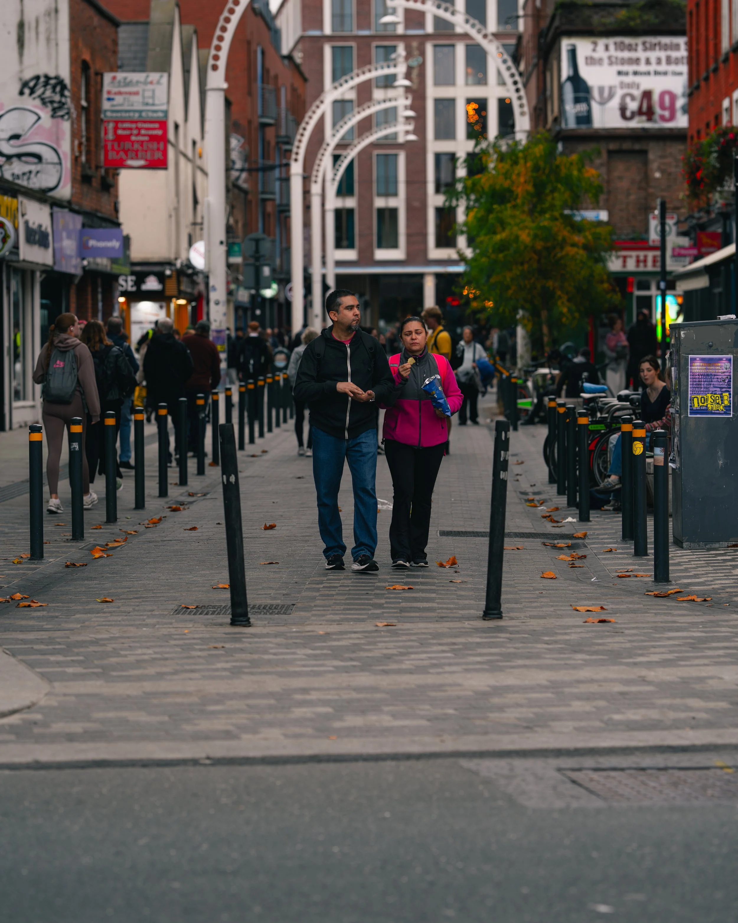 People walking on a city sidewalk with shops and signs, street lamp frames overhead, and a tree with green leaves, capturing an urban scene in the evening.