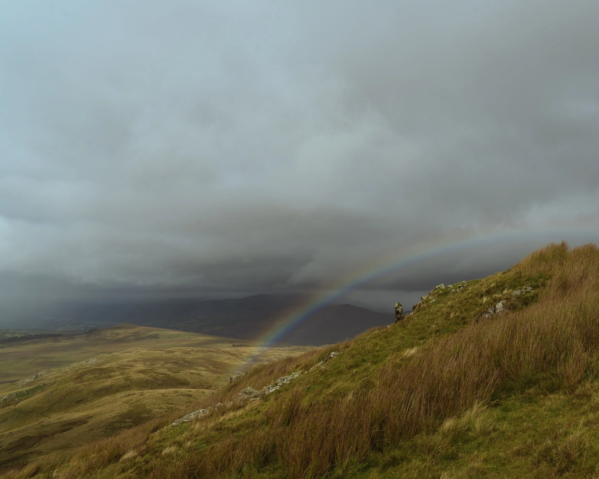 Cloudy sky over rolling grassy hills with a rainbow in the distance.