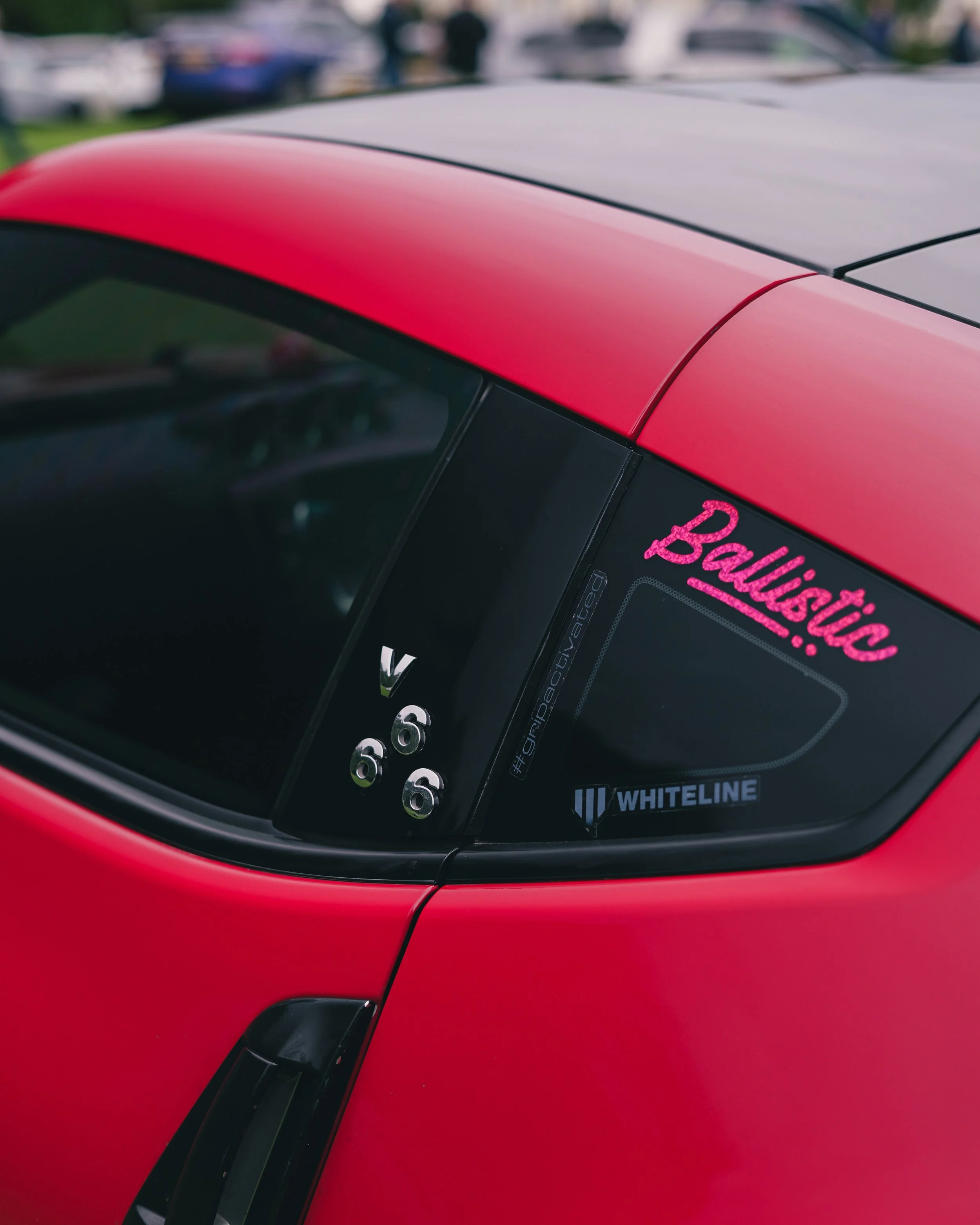 Close-up of the rear window of a red  Nissan z370 sports car with 'Ballistic' written in pink on the window, and a sticker with 'WHITELINE' on it.