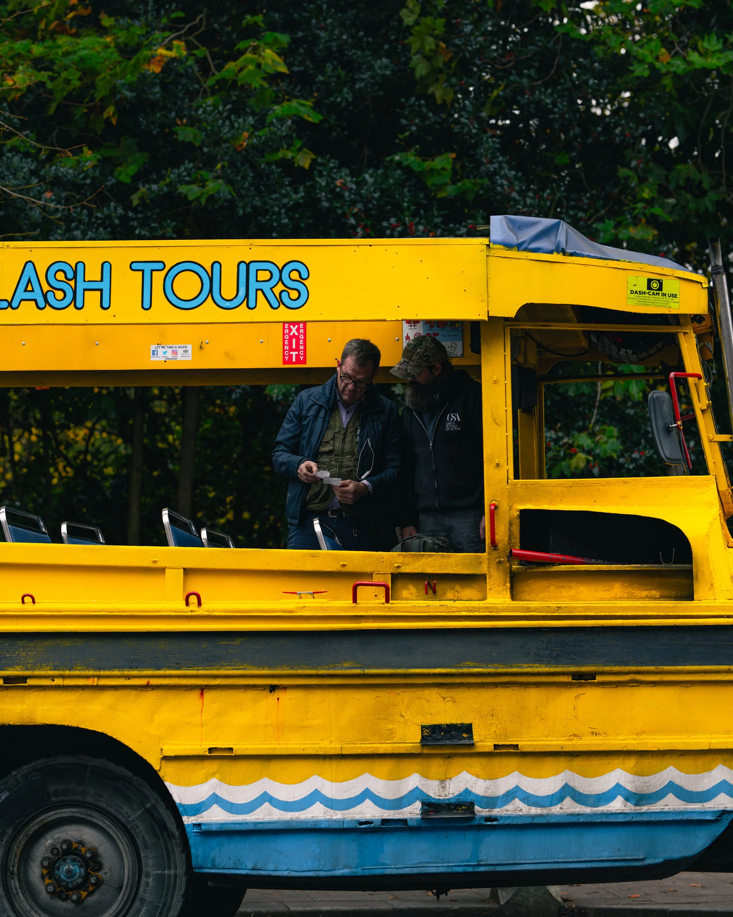 Two men standing inside a bright yellow amphibious tour bus, one showing a paper to the other, with trees in the background.