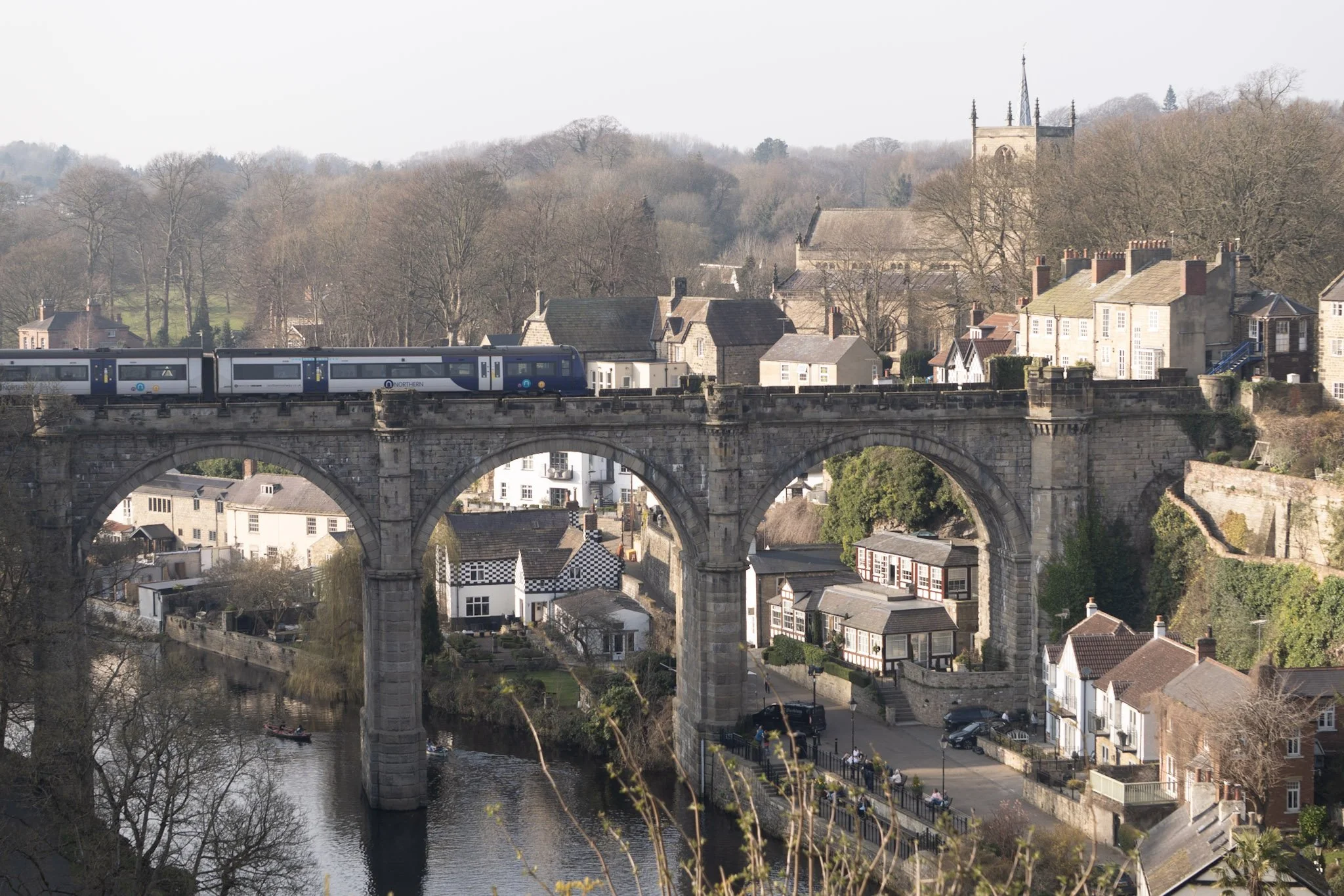 A train crossing a the Knaresborough viaduct over the river Nidd in a town with houses, a church, and wooded hills in the background.