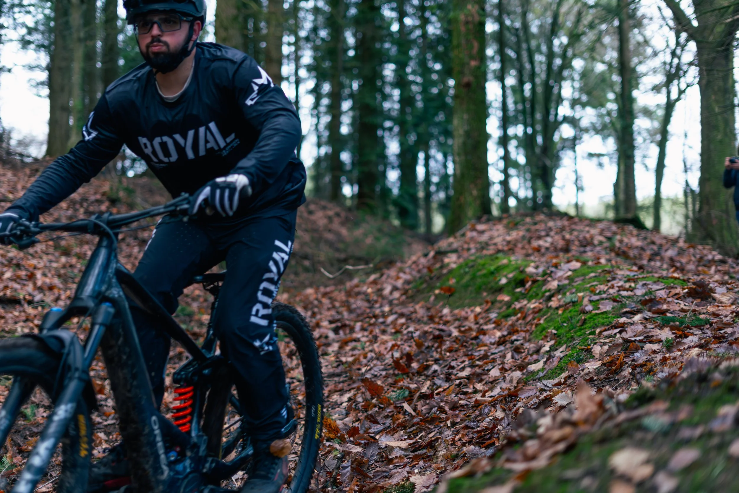 A man riding a mountain bike through a forest with fallen leaves on the ground.