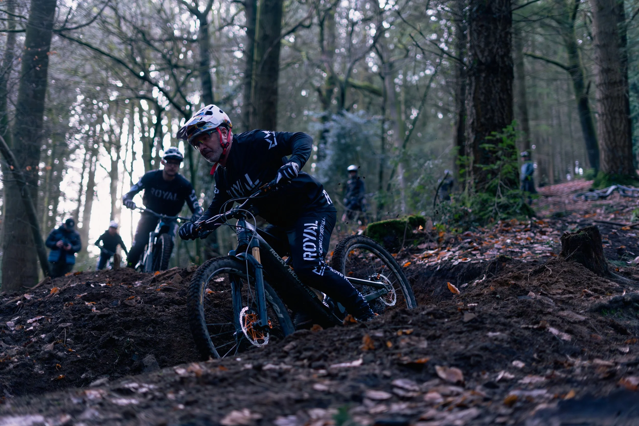 A man mountain biking on a dirt trail in a forest, surrounded by tall trees and leaf debris, with others watching in the background.