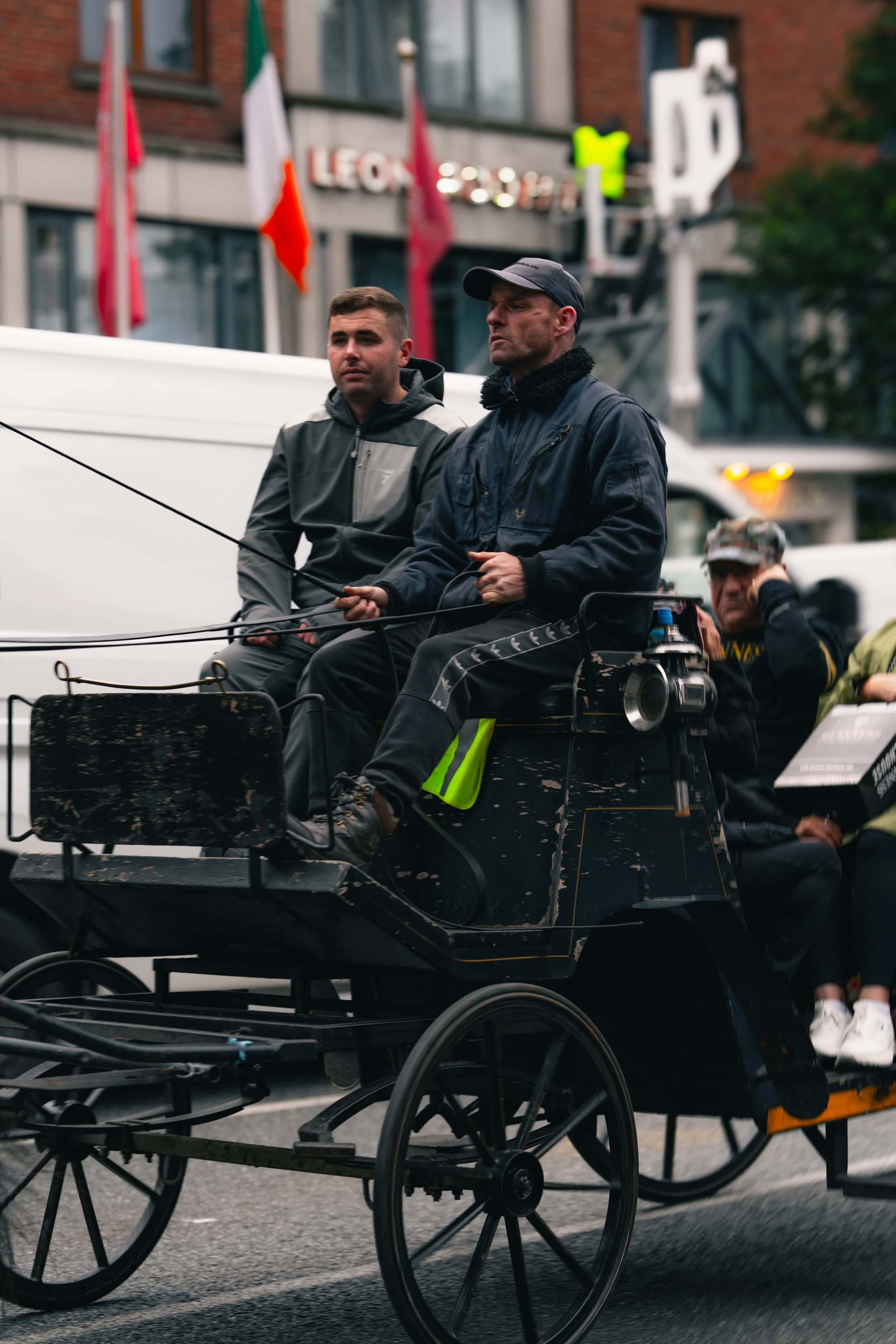 Two men riding a horse-drawn carriage in an urban street, with flags and buildings in the background.