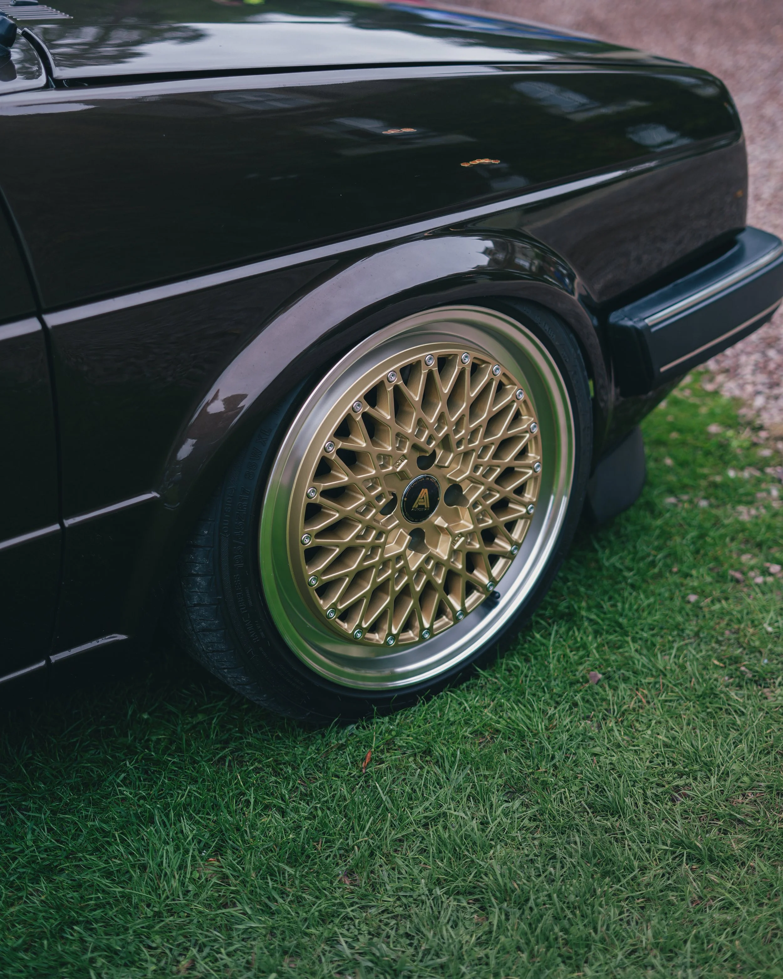 Close-up of a vintage Volkswagen Golf black car with gold custom alloy wheels parked on grass.