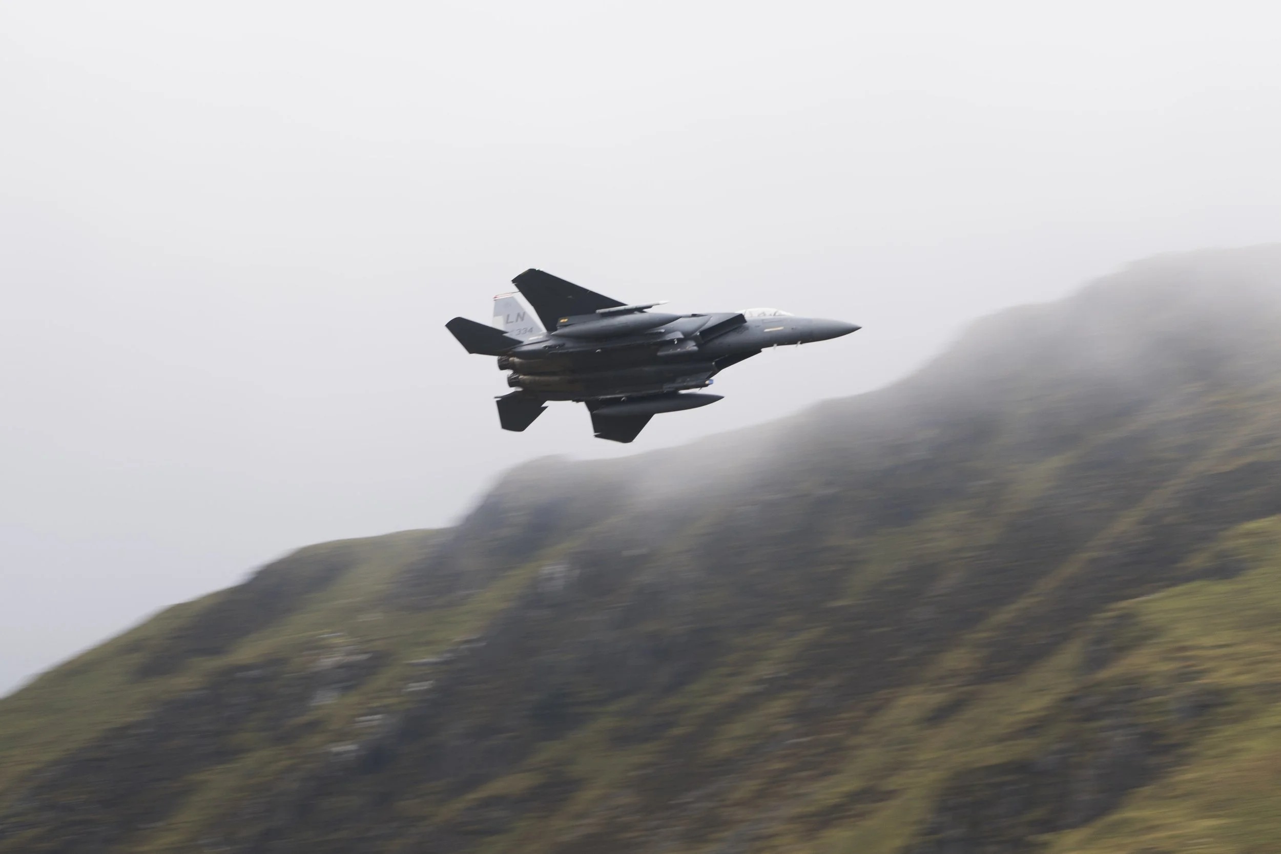 A fighter jet flying over a mountainous landscape with cloudy skies.