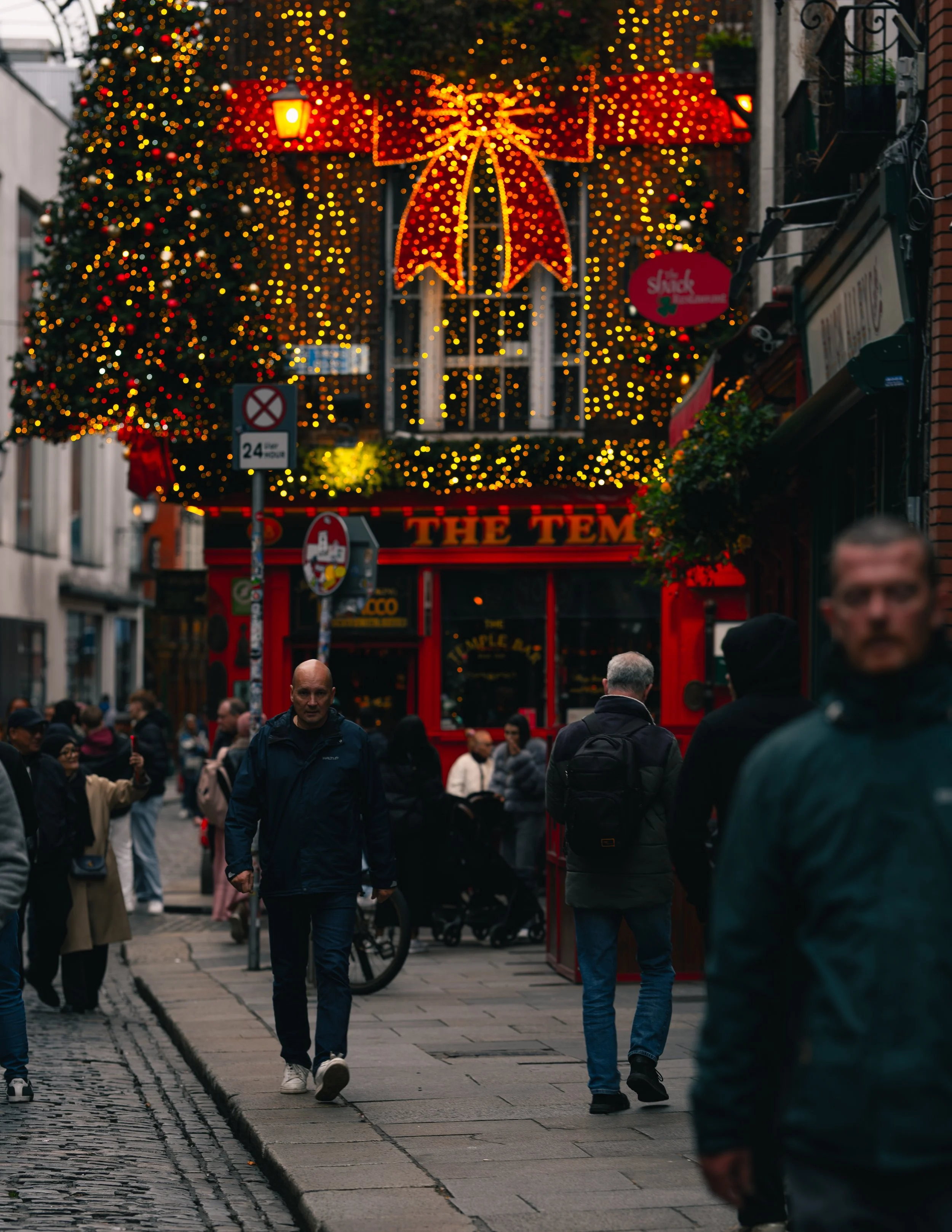 A busy city street decorated with Christmas lights and a large illuminated bow. People are walking along the sidewalk, and some are gathered outside a red storefront.
