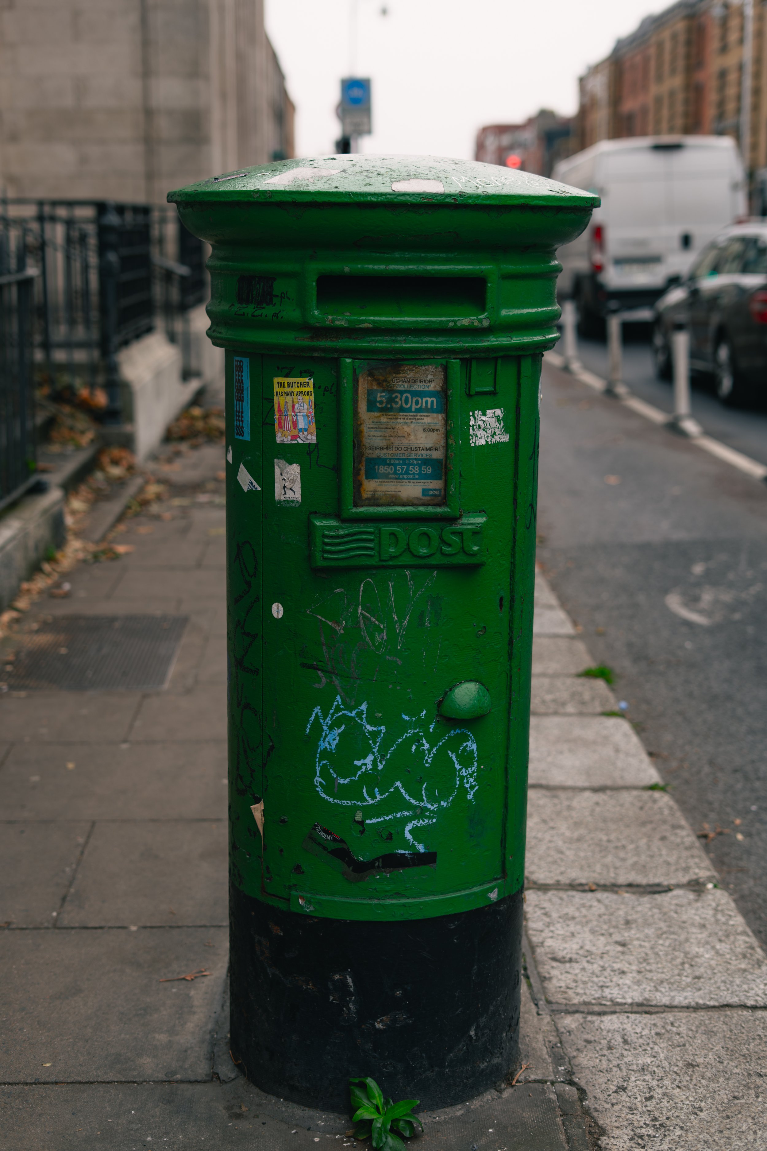 A green U.S. Postal Service mailbox on a city sidewalk with graffiti and stickers, with cars and buildings in the background.