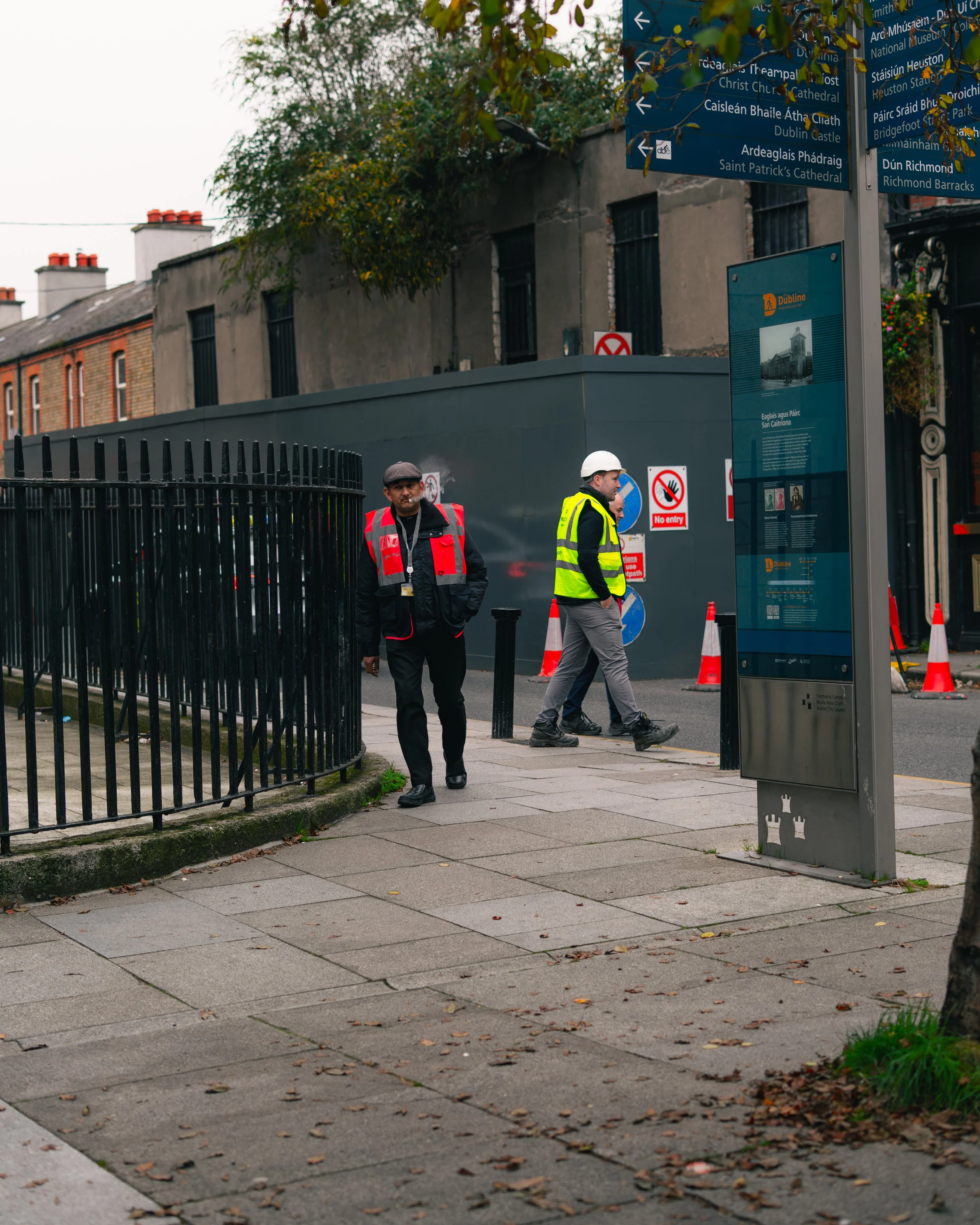 Two men in high-visibility vests and a hard hat walking on sidewalk near construction site with orange cones, signs, and information board, in an urban area with multi-story buildings and overcast sky.