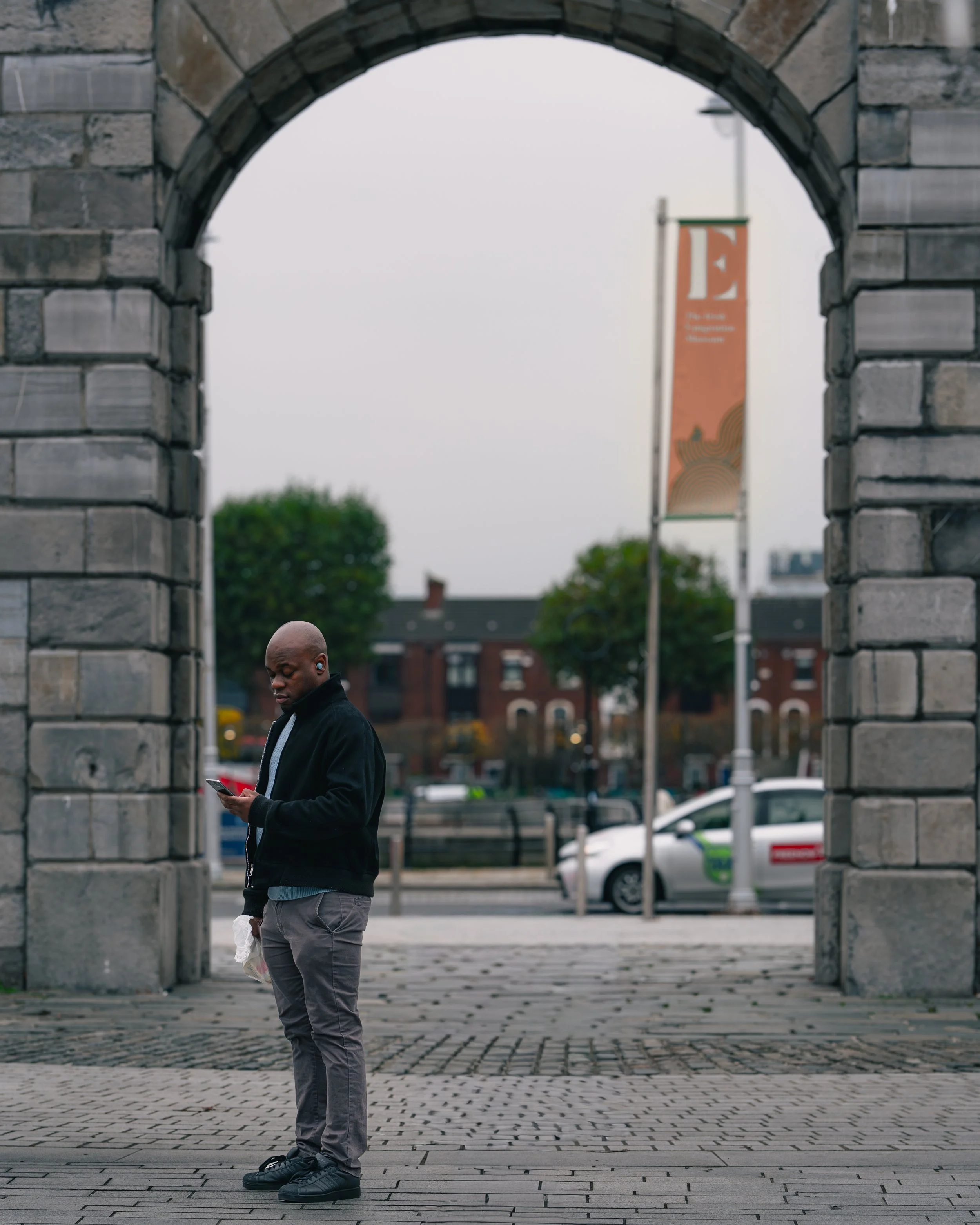 A man standing under an arched stone structure, looking at his phone, with cars and trees in the background.