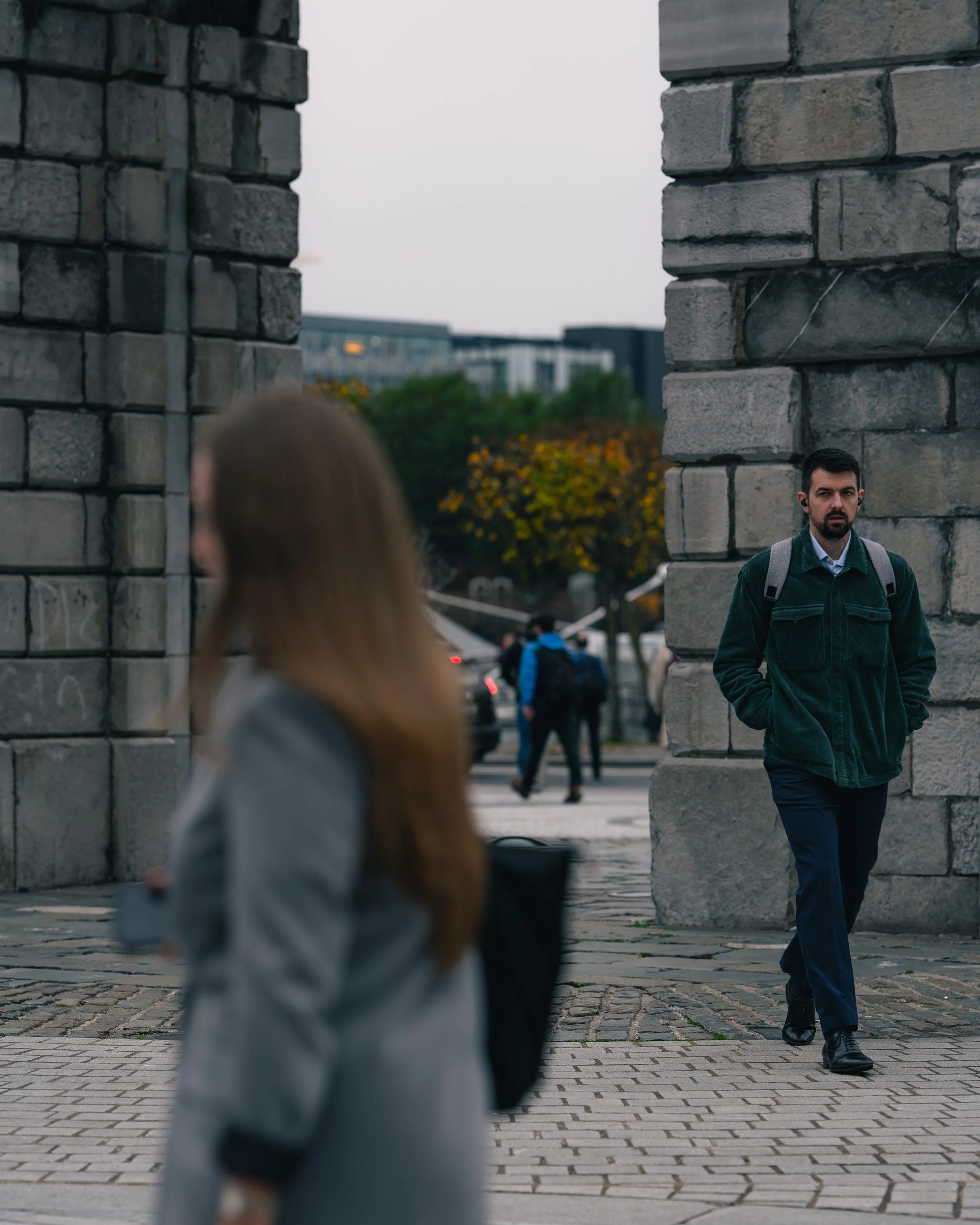 A man in a green jacket and blue pants walking on a sidewalk with a backpack, passing between large stone pillars. In the foreground, a woman with long hair, blurred, is holding a drink and a black bag, walking in the opposite direction. In the backg