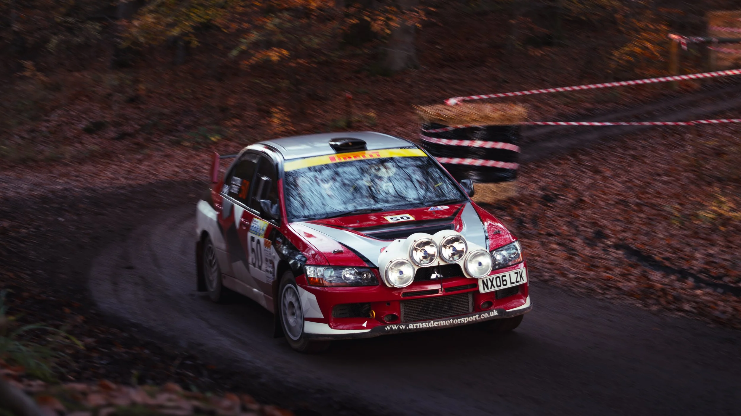 Rally car racing on a dirt road through a wooded area during autumn, with leaves on the ground and a red and white striped barrier on the side. wye dean rally 