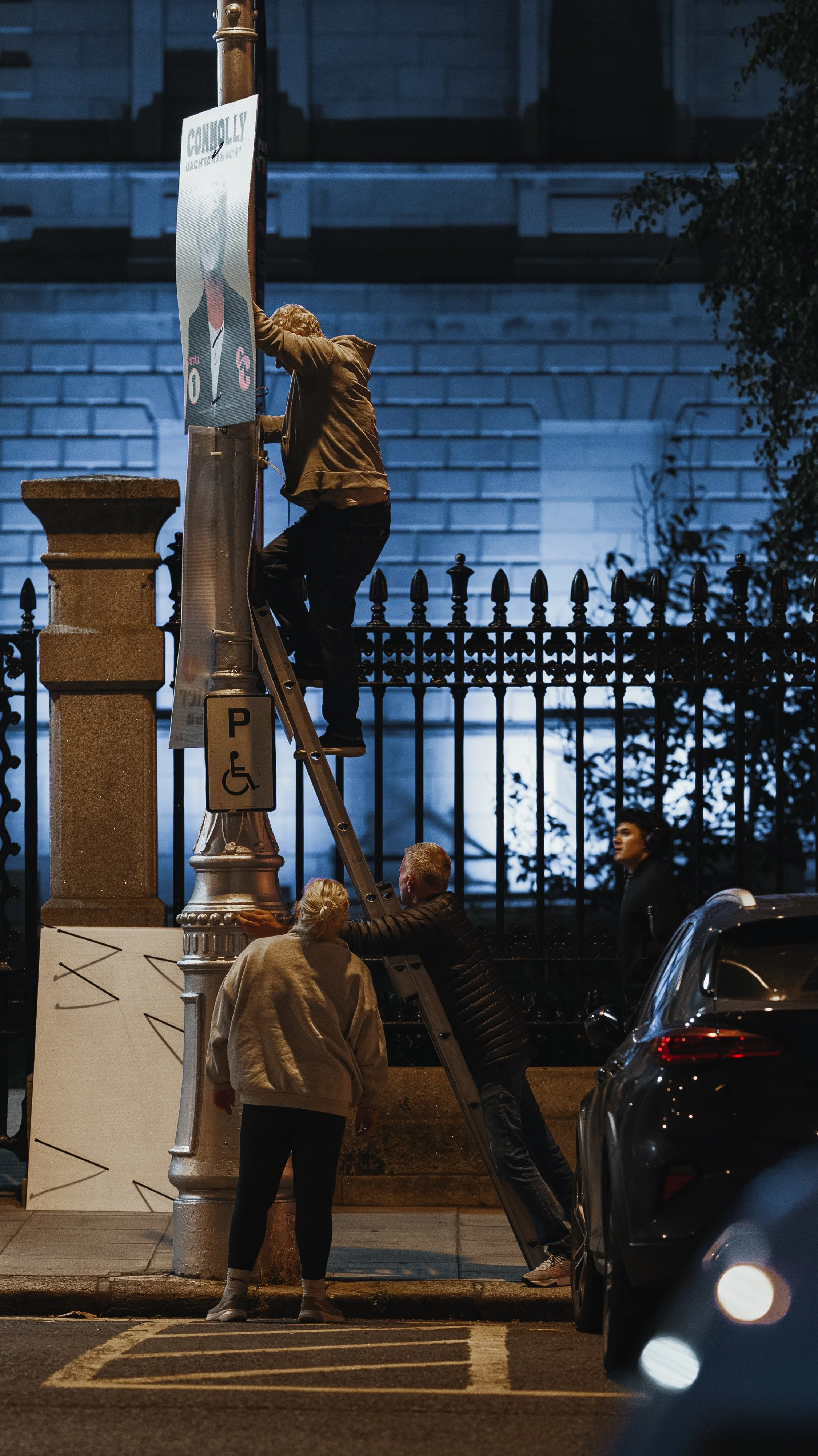 People installing or repairing a billboard on a street at night, with a car parked nearby and a fence in the background.