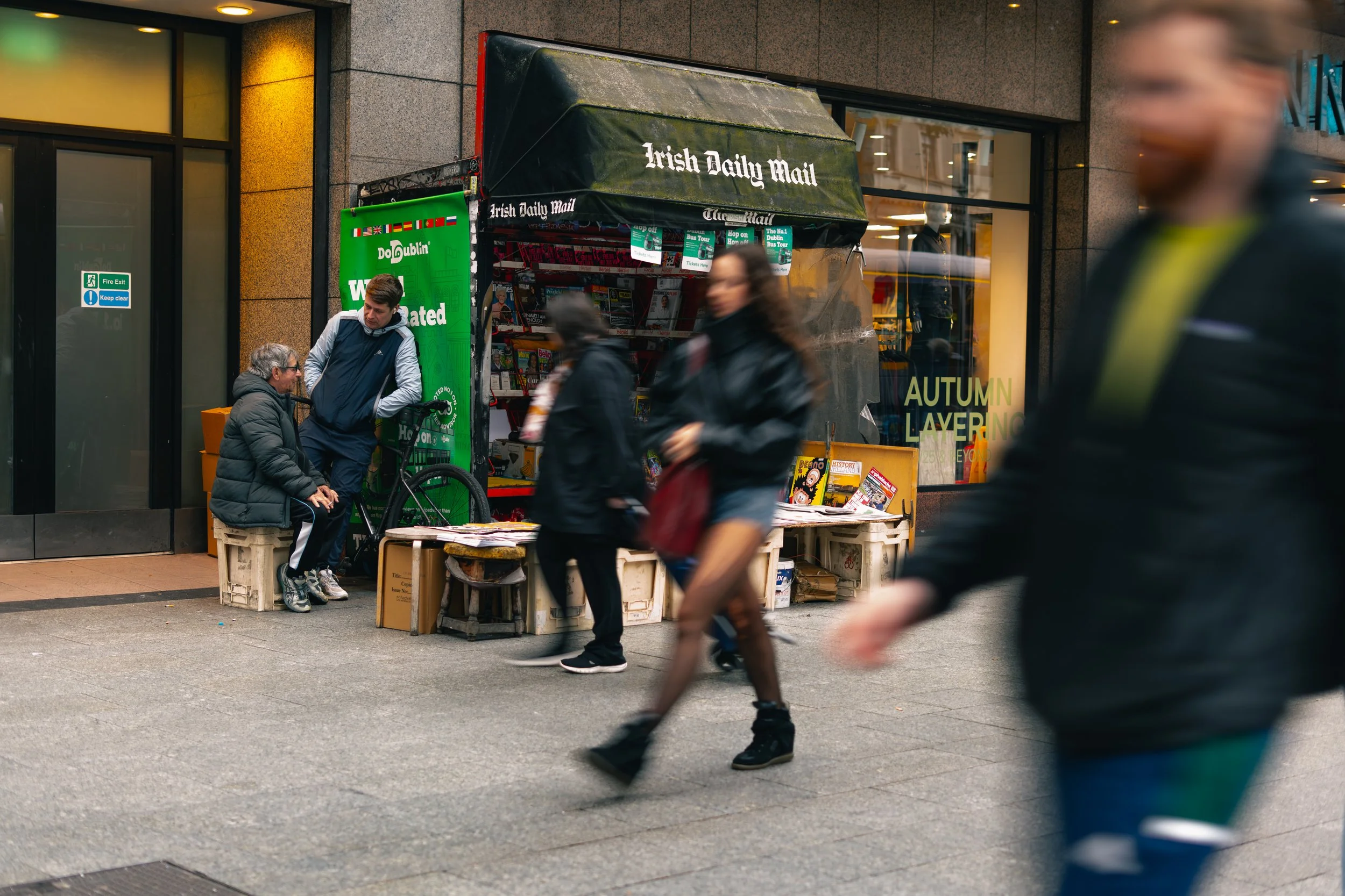 People walking past a newsstand in an urban setting, with an Irish Daily Mail kiosk and a display of magazines and newspapers outside.
