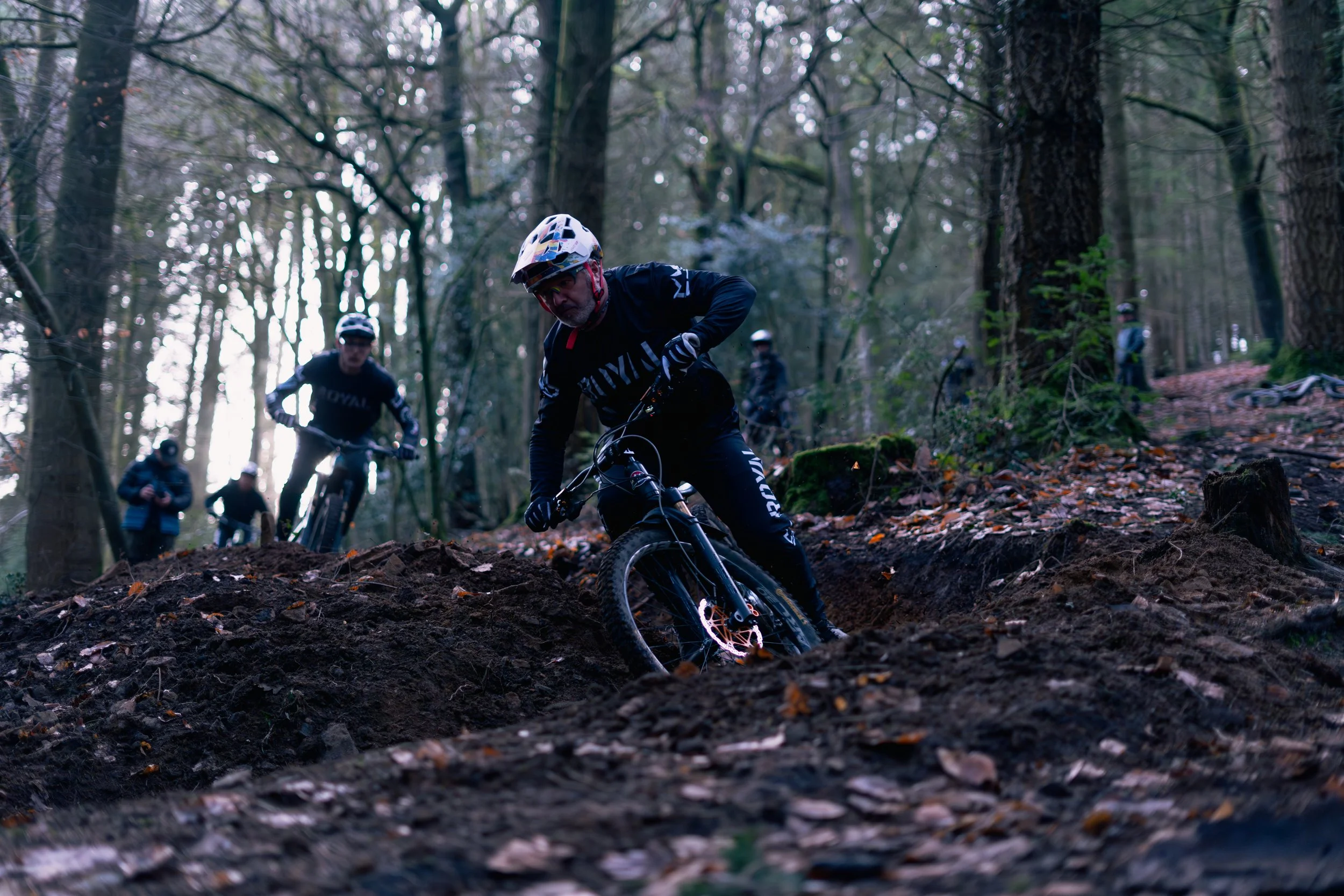 A group of mountain bikers riding through a forest trail with tall trees and fallen leaves, in a downhill course.