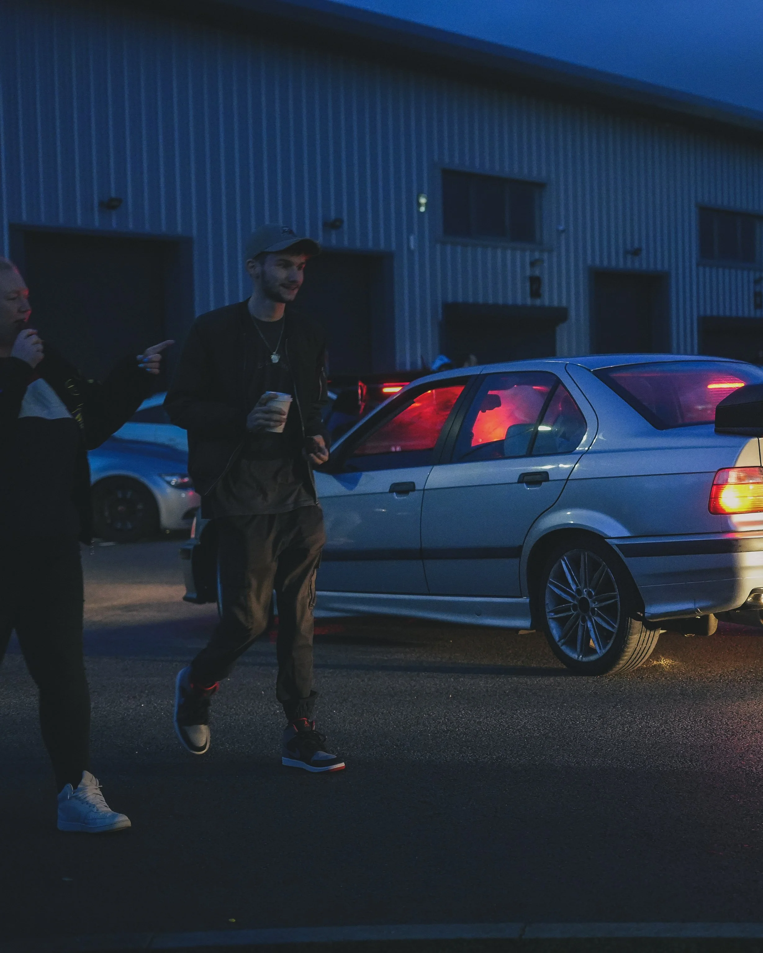 Two young men walking outside at night near parked cars, one holding a drink, with a warehouse in the background.
