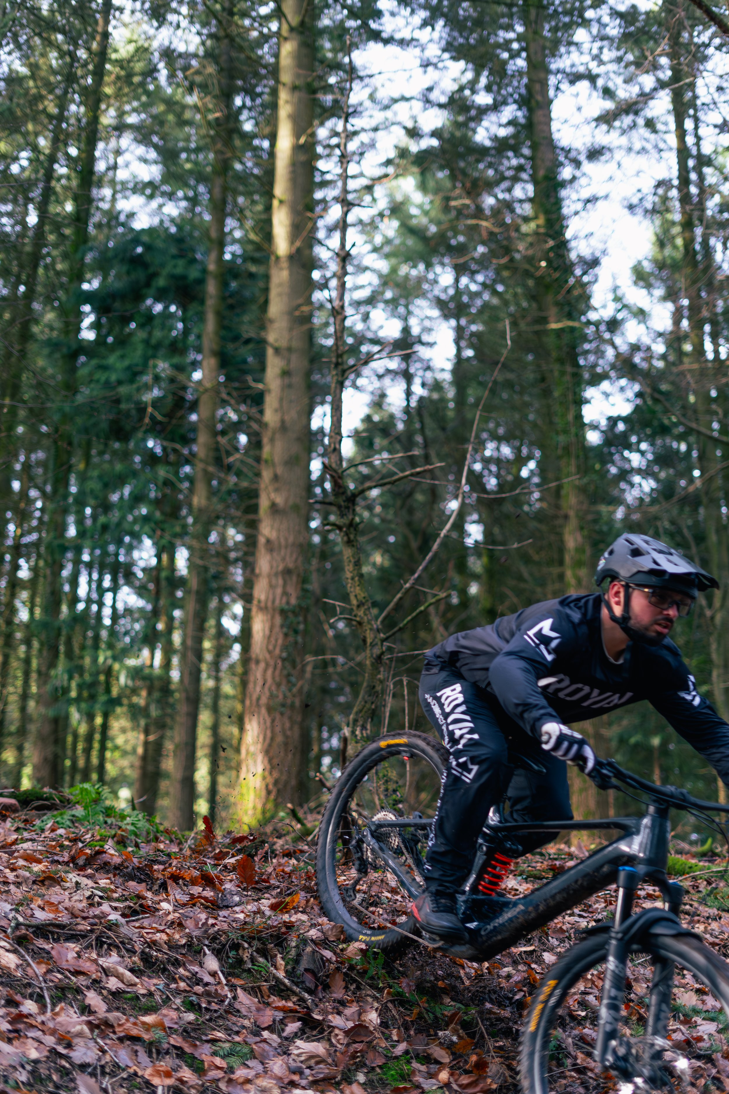 A man riding a mountain bike downhill through a forested trail covered with fallen leaves.