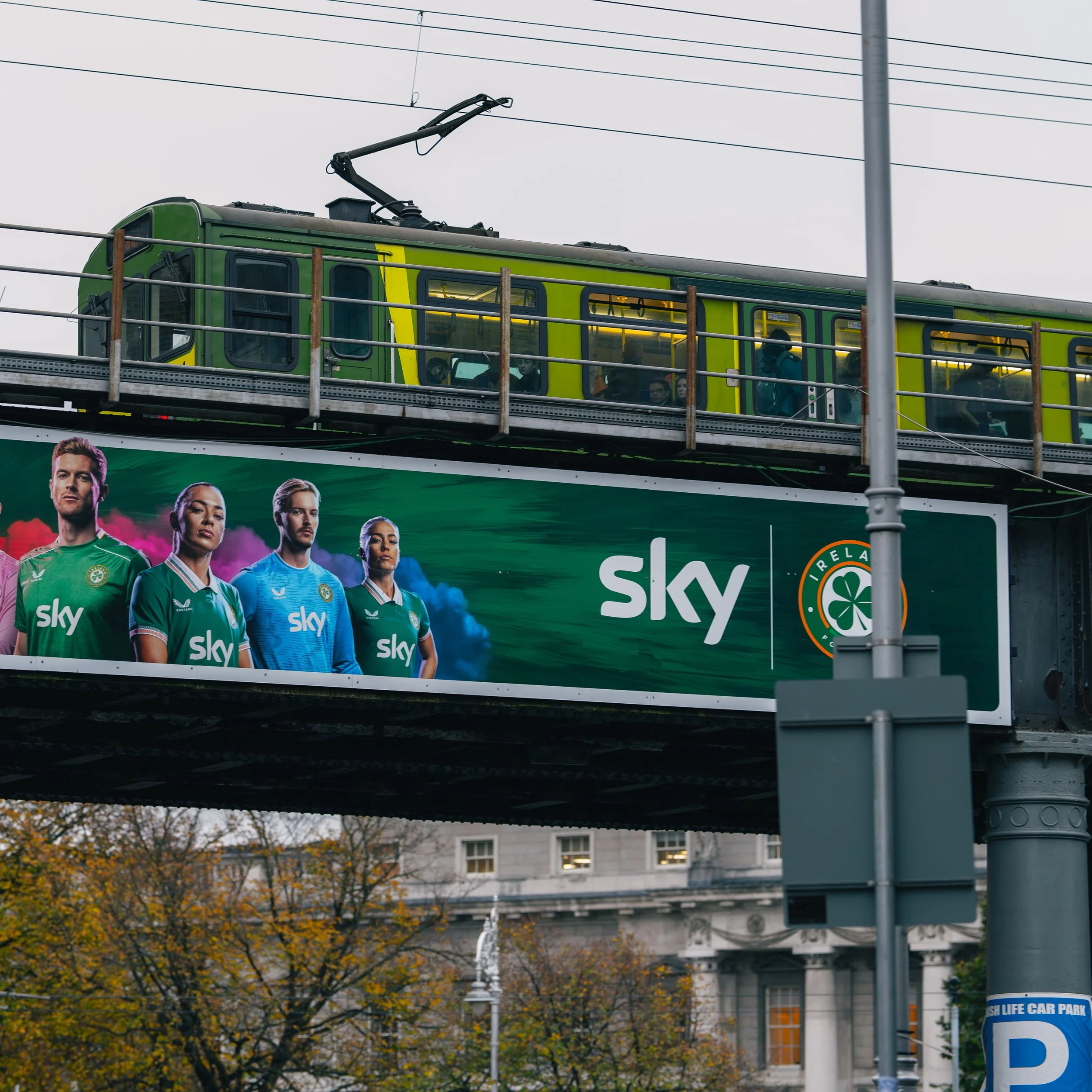 An elevated train with a green exterior runs above a street. Beneath the train is a large billboard advertising Sky sports, featuring four athletes in green and blue sports jerseys, with a cityscape and trees in the background.
