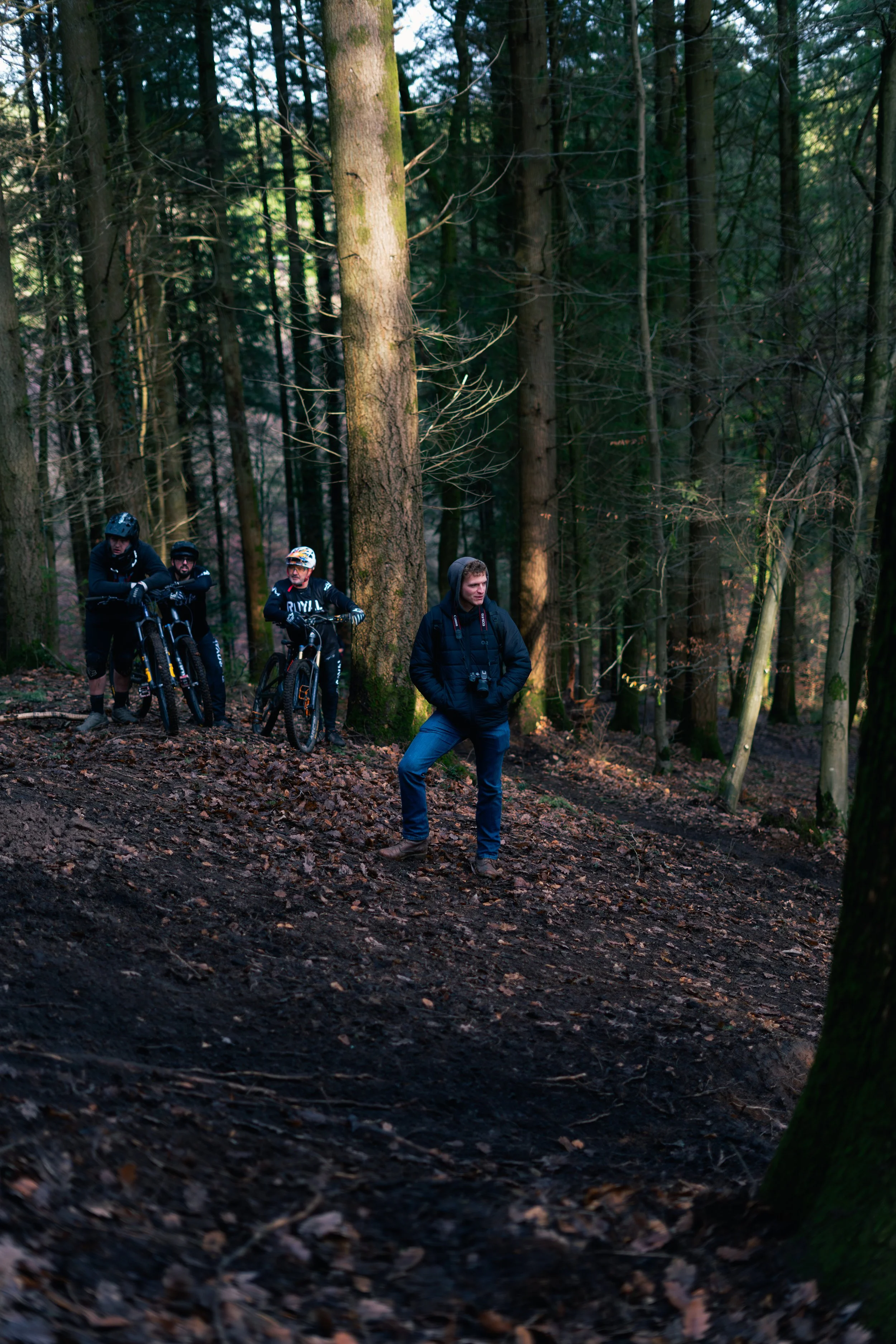 A group of four mountain bikers resting in a forested area, with one person standing and three others with bikes, all dressed in outdoor gear.