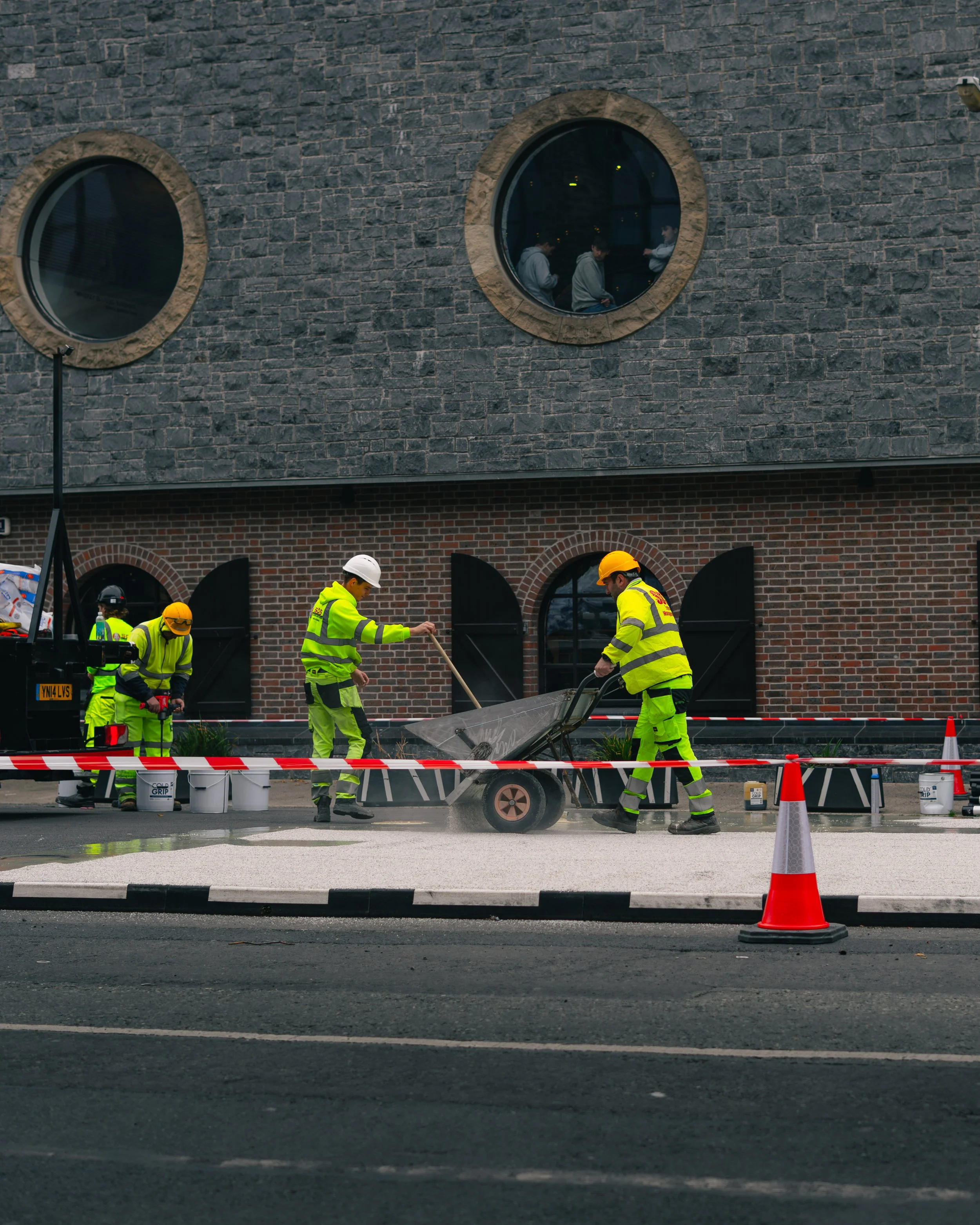Construction workers in bright yellow safety gear working on road pavement, with some inside a fenced-off area and others pushing a wheelbarrow, in front of a brick building with round windows.