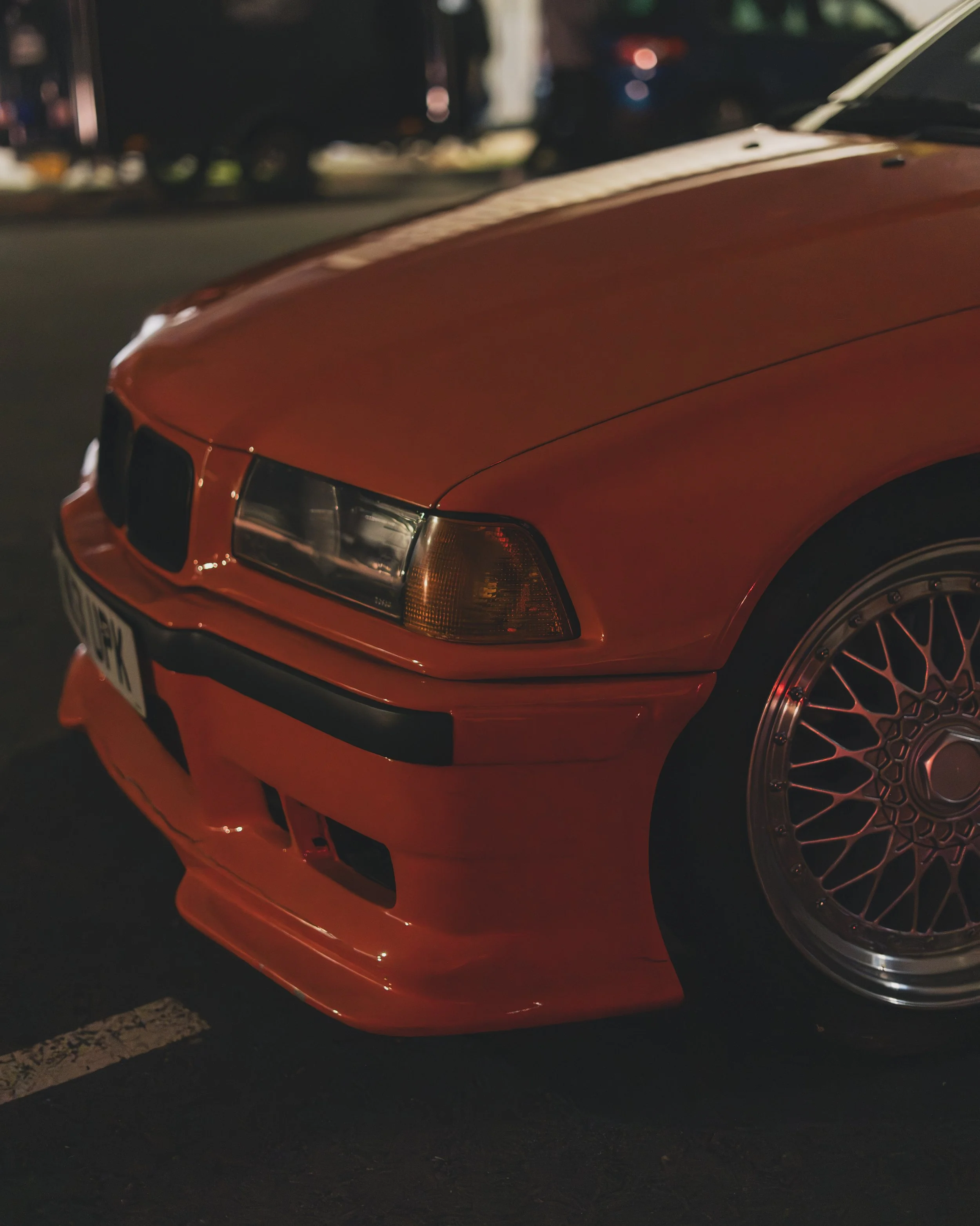 Close-up of the front part of a red vintage BMW car with a black bumper, parked in a low-lit area at night.