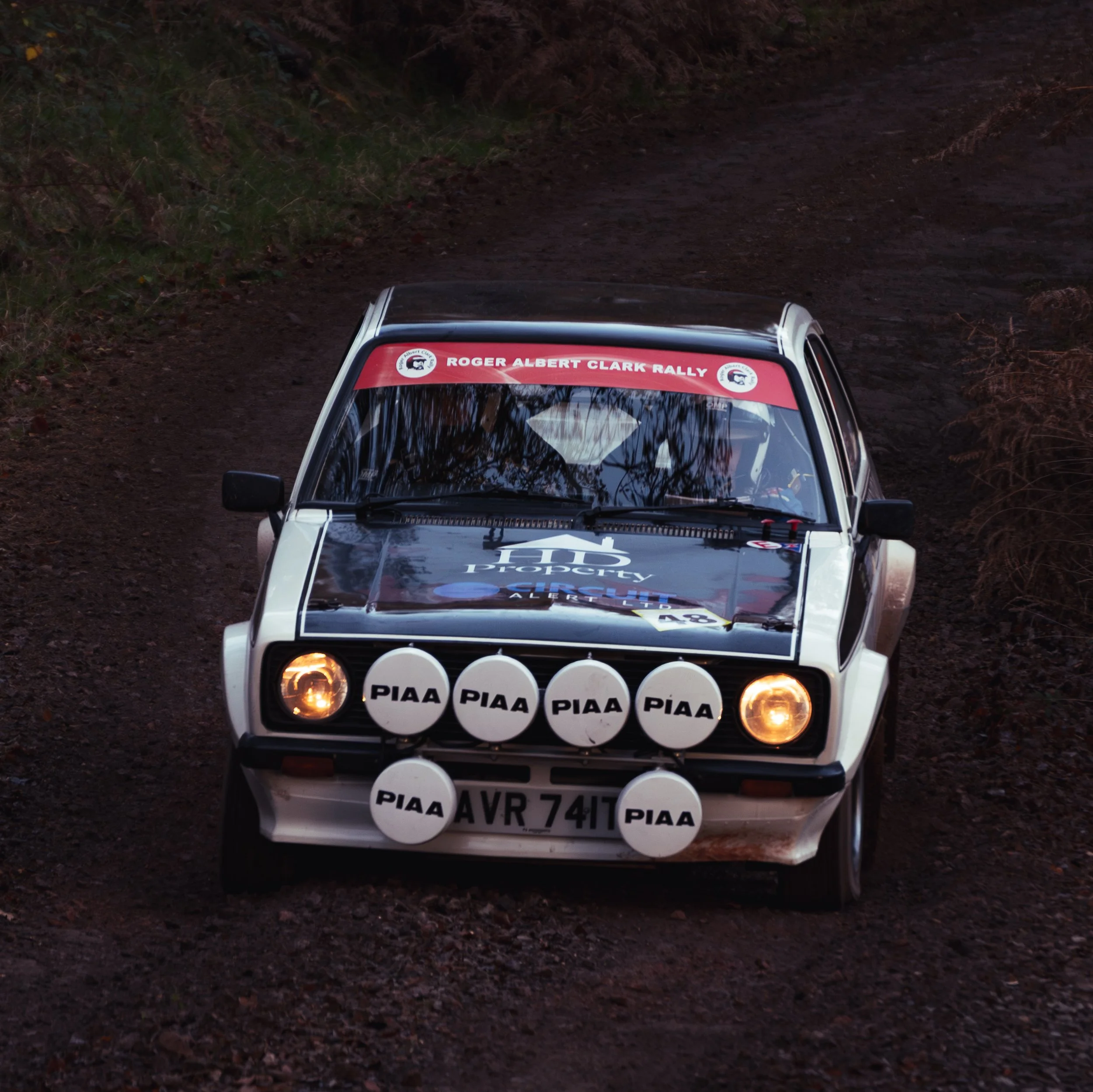 A white rally car with multiple PIAA auxiliary lights on the front, driving on a dirt trail in a wooded area during twilight or dusk, with a red banner on the windshield reading 'Roger Albert Clark Rally.'