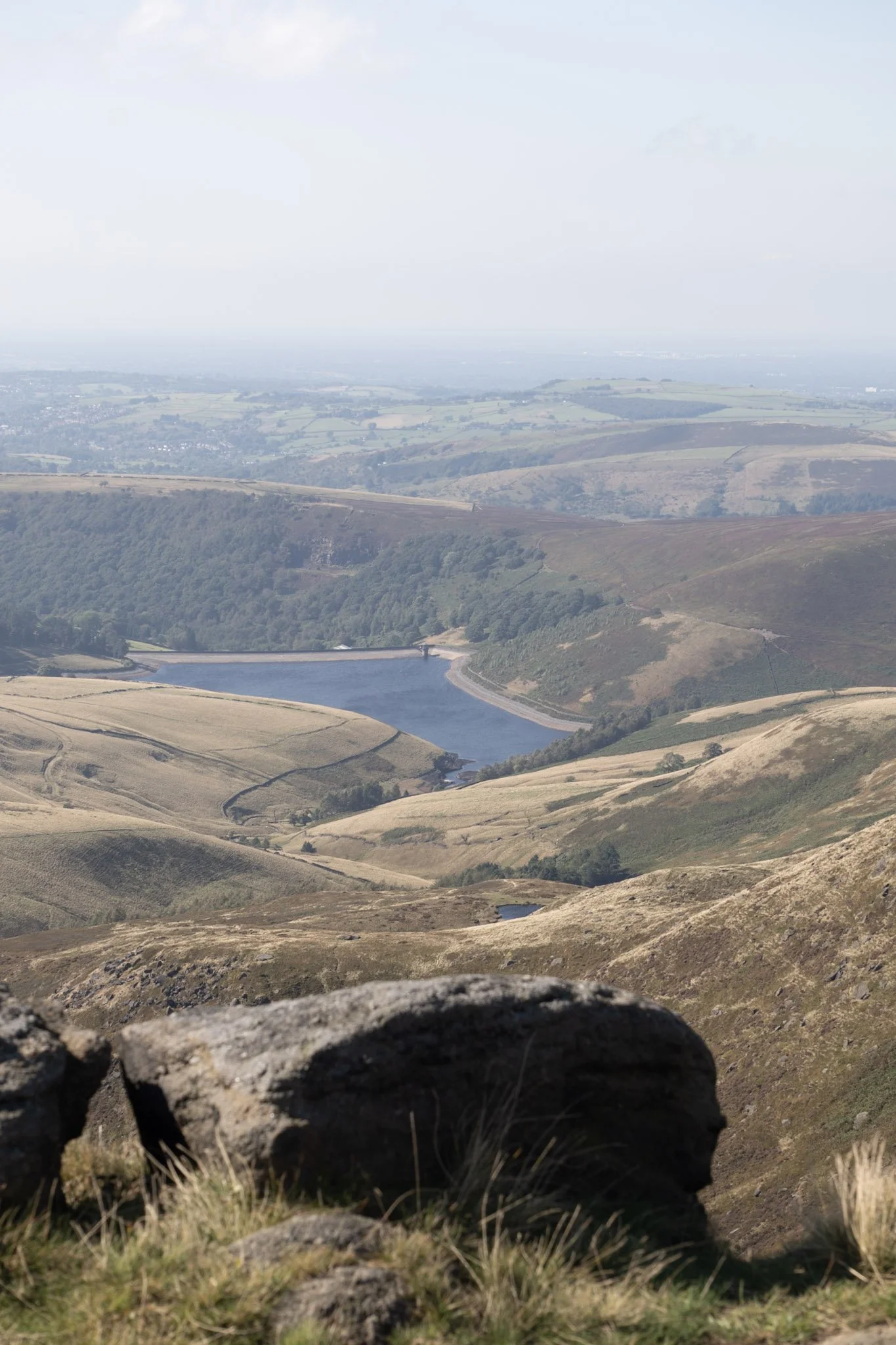 Hilly landscape with a reservoir or lake, rolling grassy hills, scattered trees, and a stone in the foreground, under a cloudy sky.