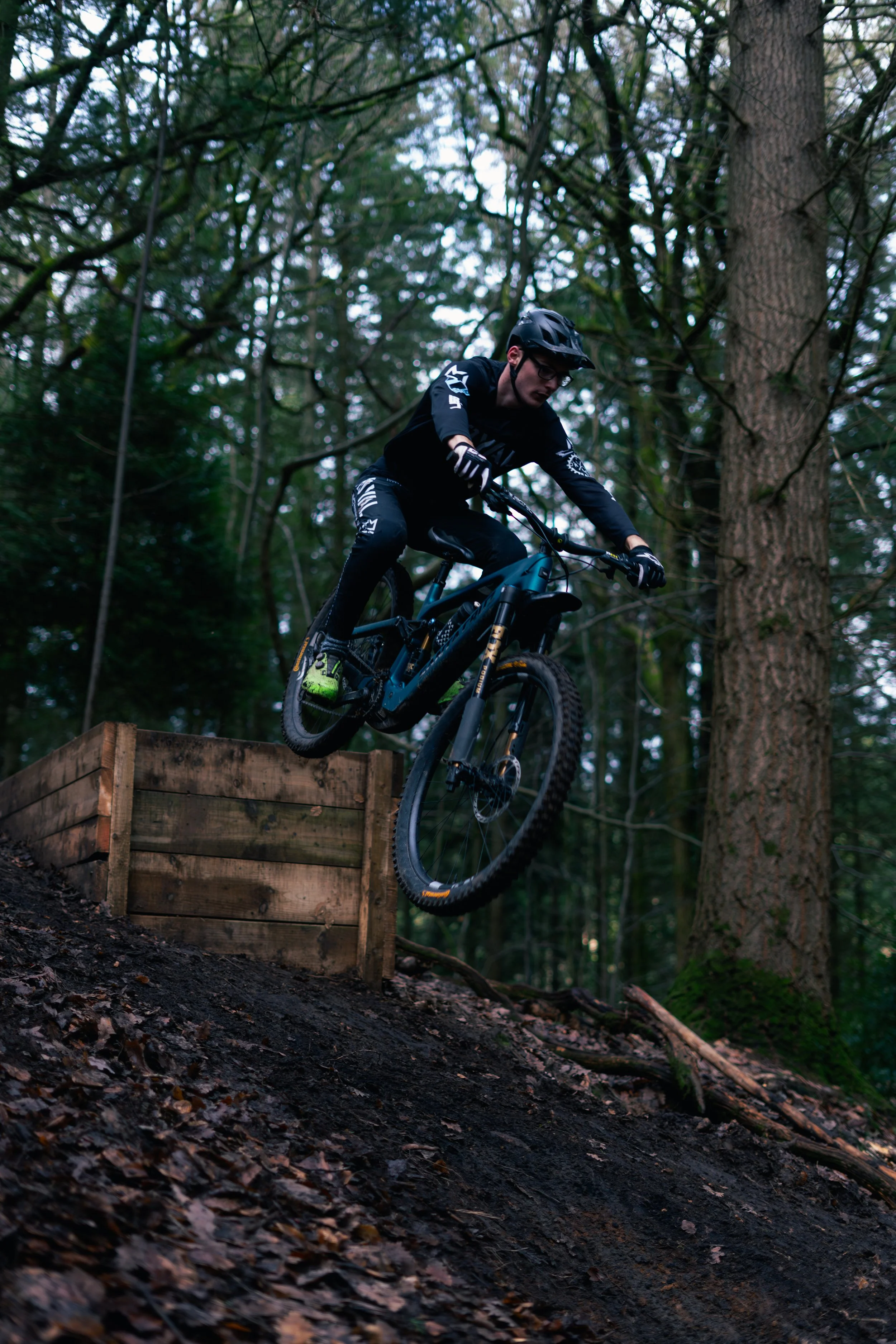 Mountain biker wearing black gear and helmet jumping off a wooden ramp in a forested area.