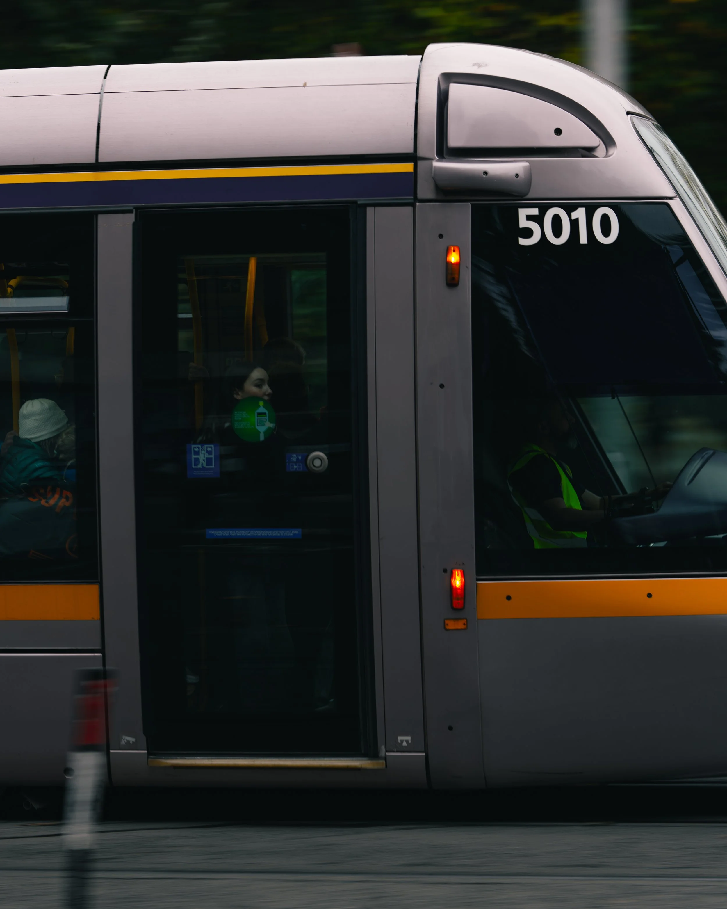 A modern gray and yellow city bus with the number 5010 on the front, moving along the road with a driver in a safety vest and passengers visible inside.