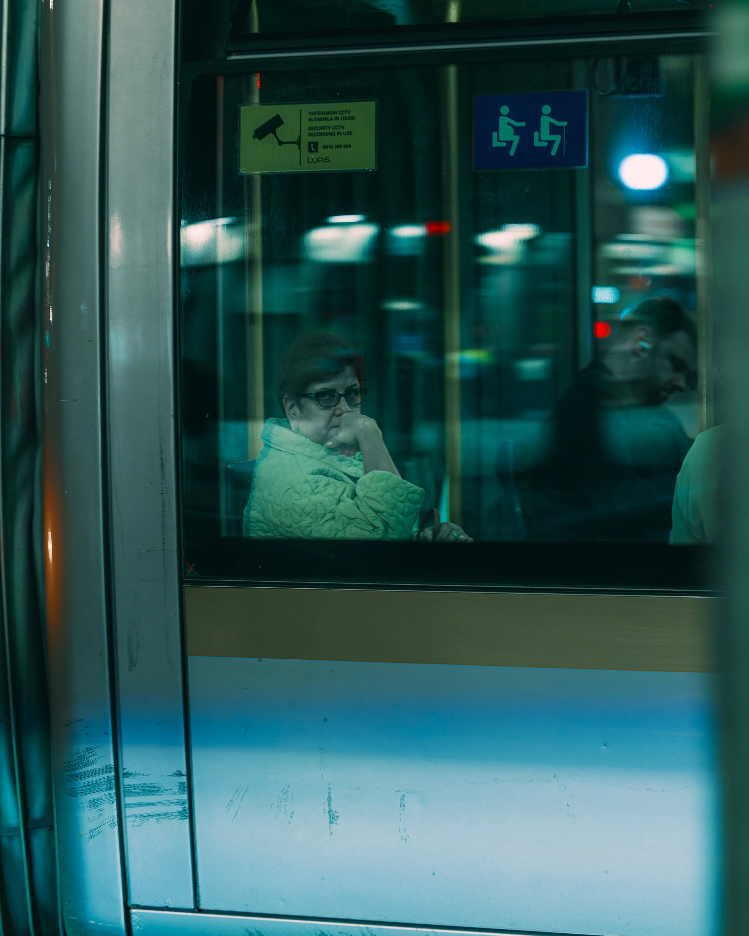 Woman sitting on a bus or train, wearing glasses and a yellow quilted jacket, looking through the window with another passenger in the background.