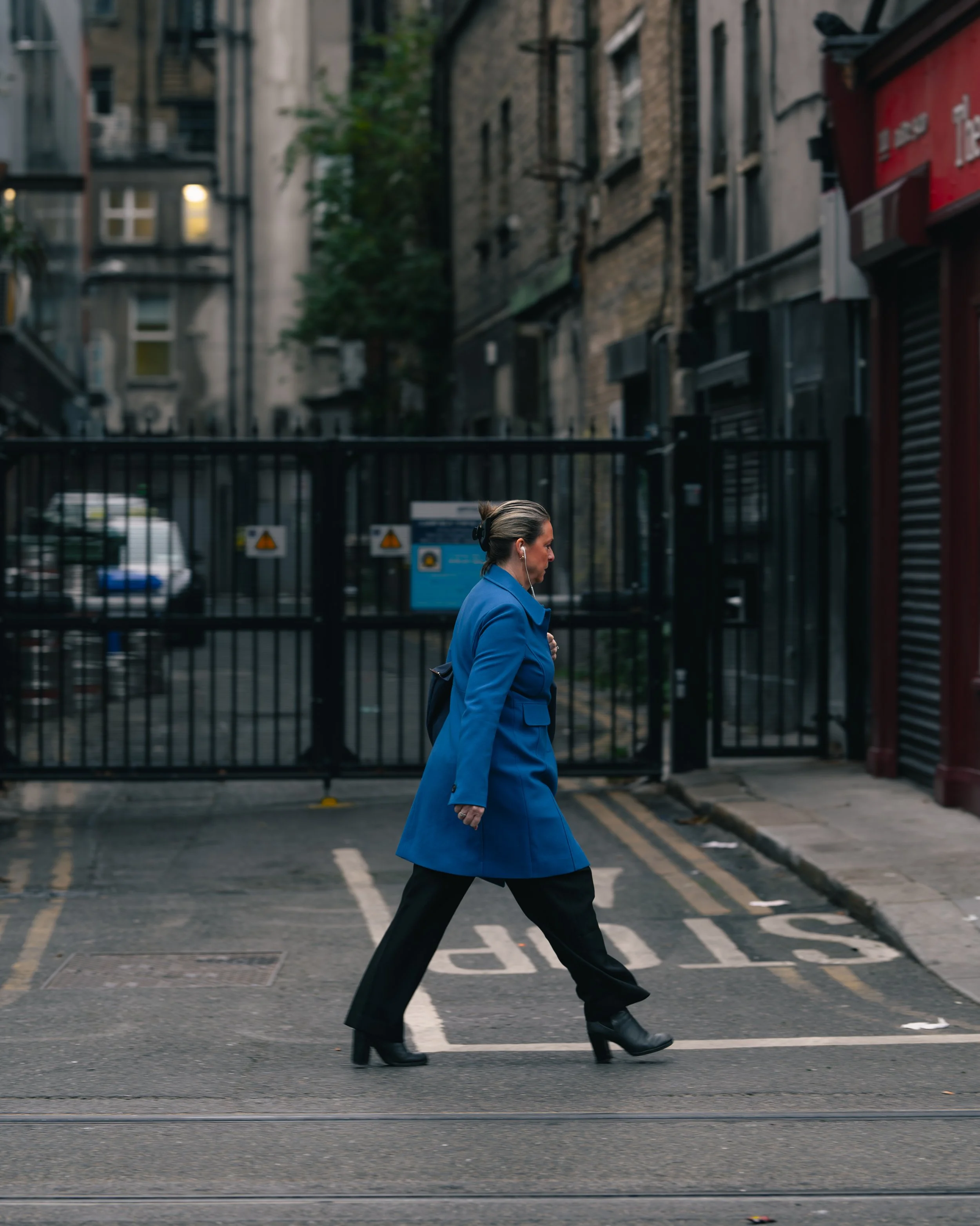 A woman in a blue coat and high heels walking across a city street at an intersection.