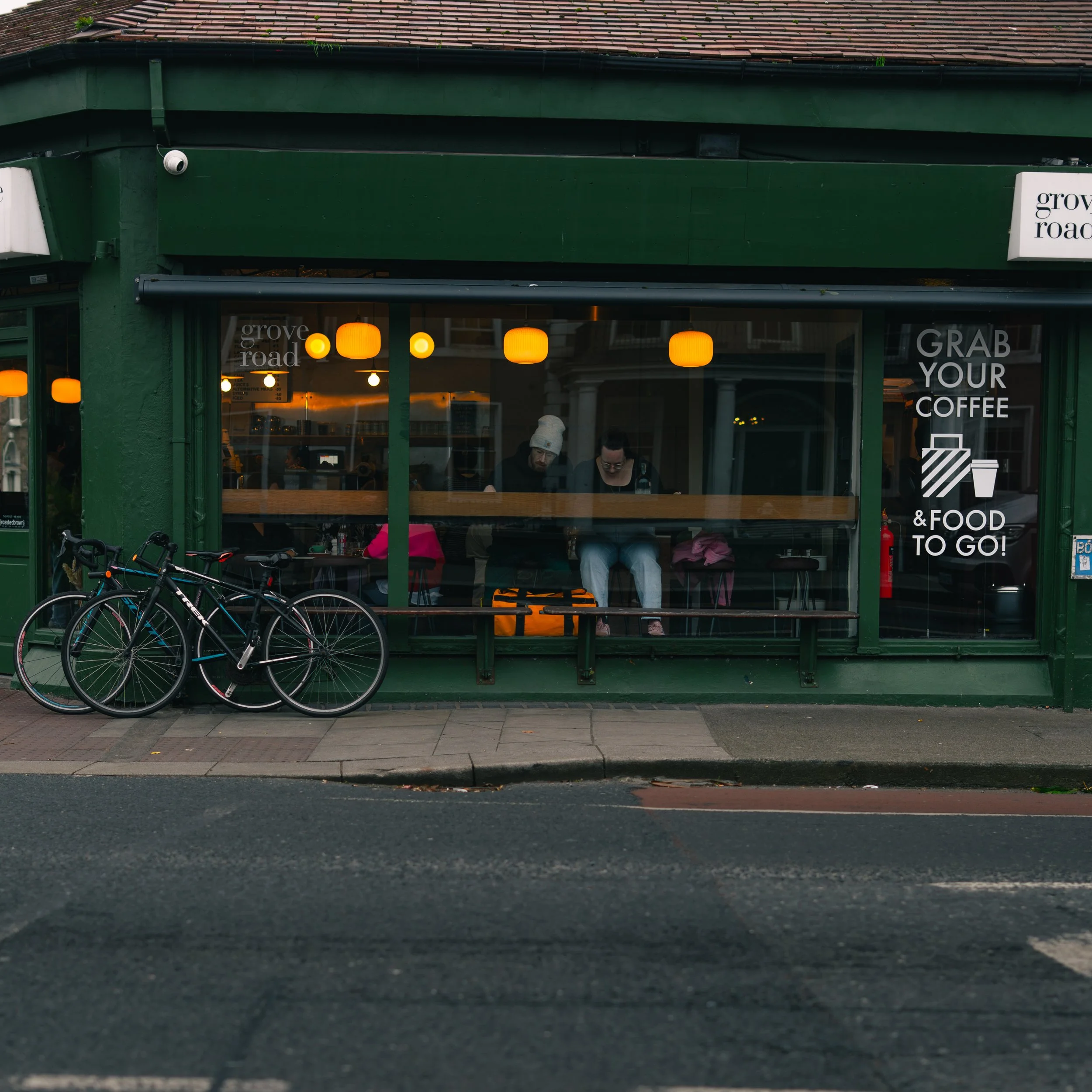 A view of a green storefront with large windows showing two people seated inside at a coffee shop. The window has signage that reads 'GRAB YOUR COFFEE & FOOD TO GO!' Below the window, three bicycles are parked on the sidewalk. The interior has warm l