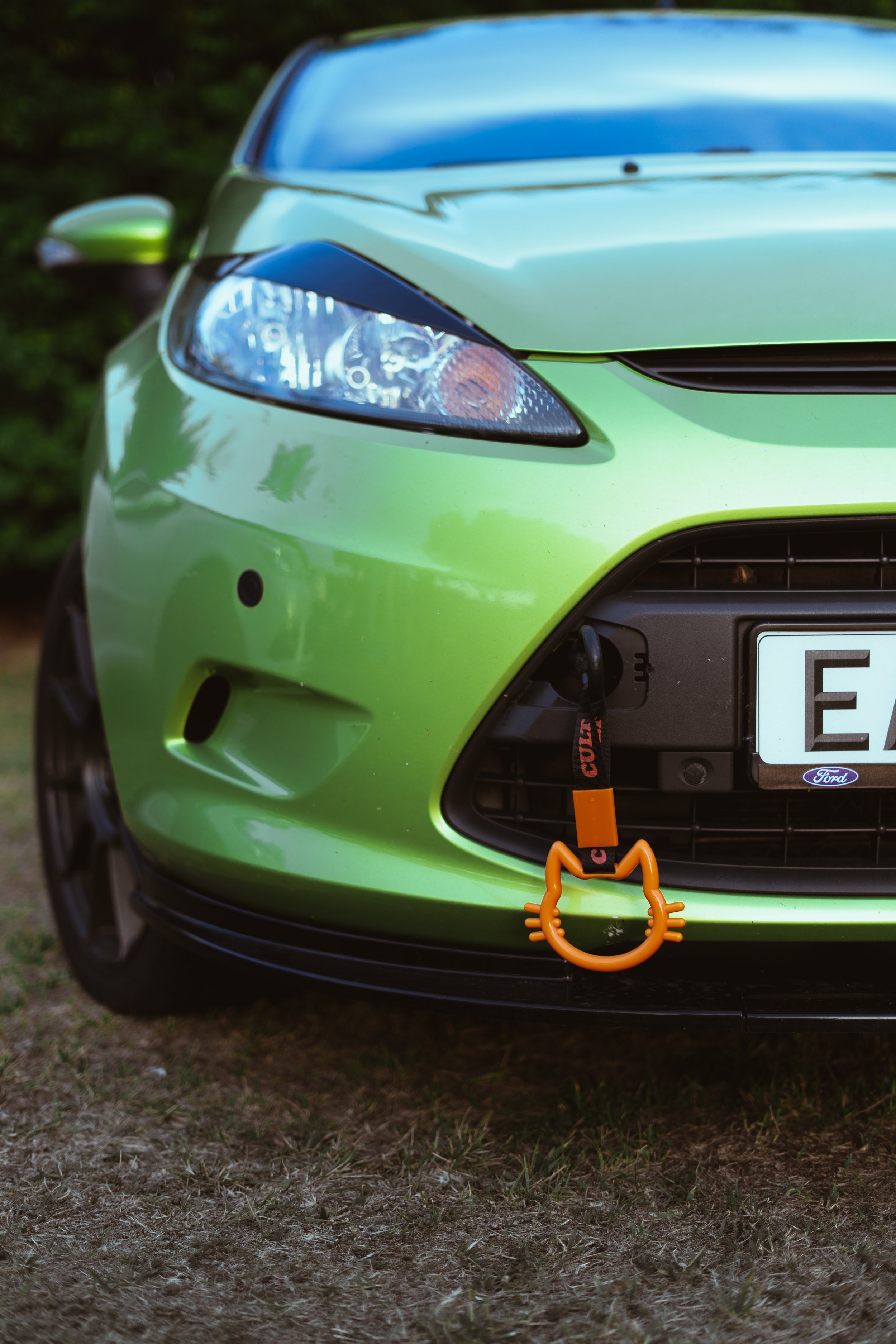 Close-up of the front of a bright green Ford Festa car with a cat-shaped orange keychain hanging from the front grille.