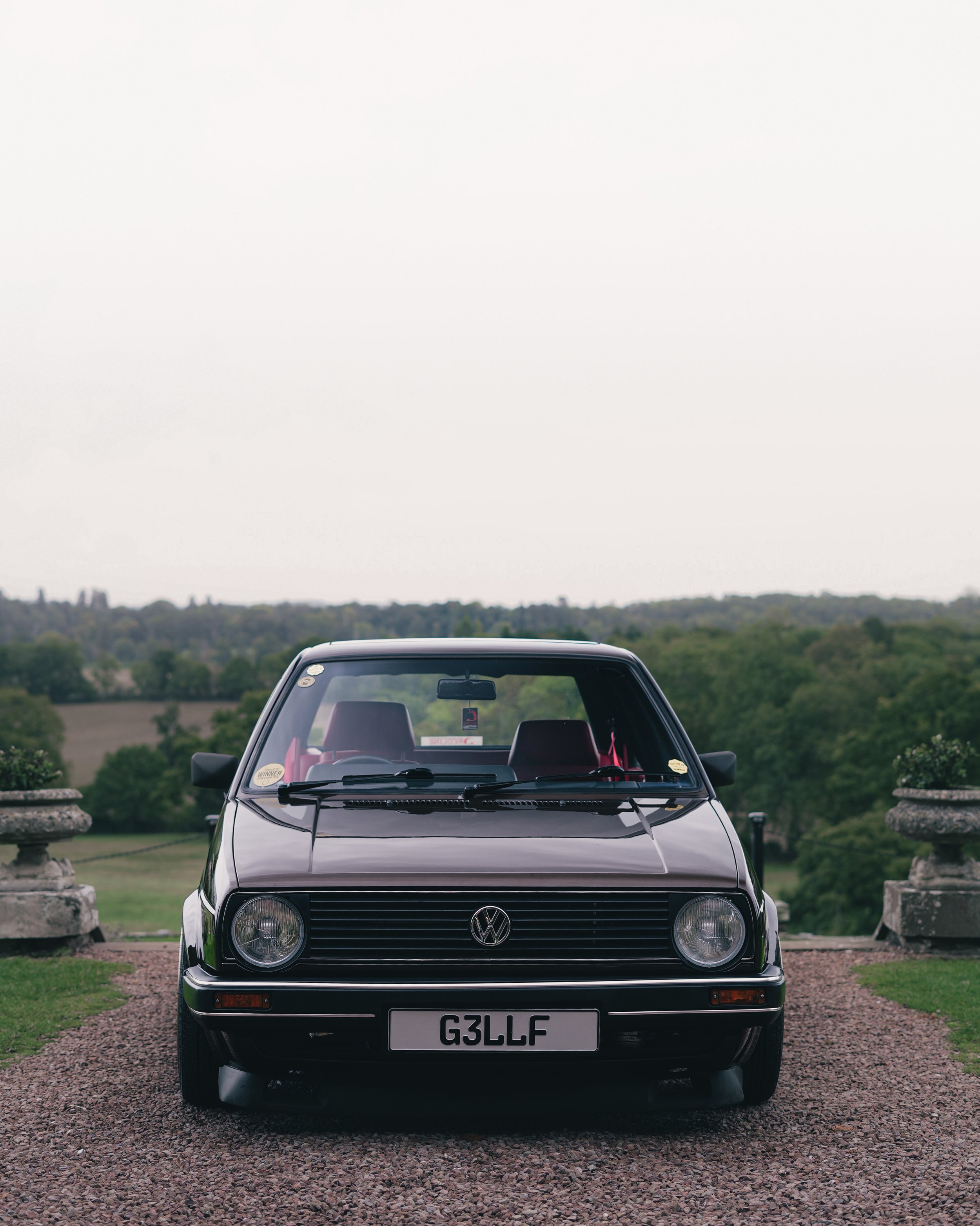 Front view of a black vintage Volkswagen Golf parked on a gravel pathway with green trees and hills in the background.