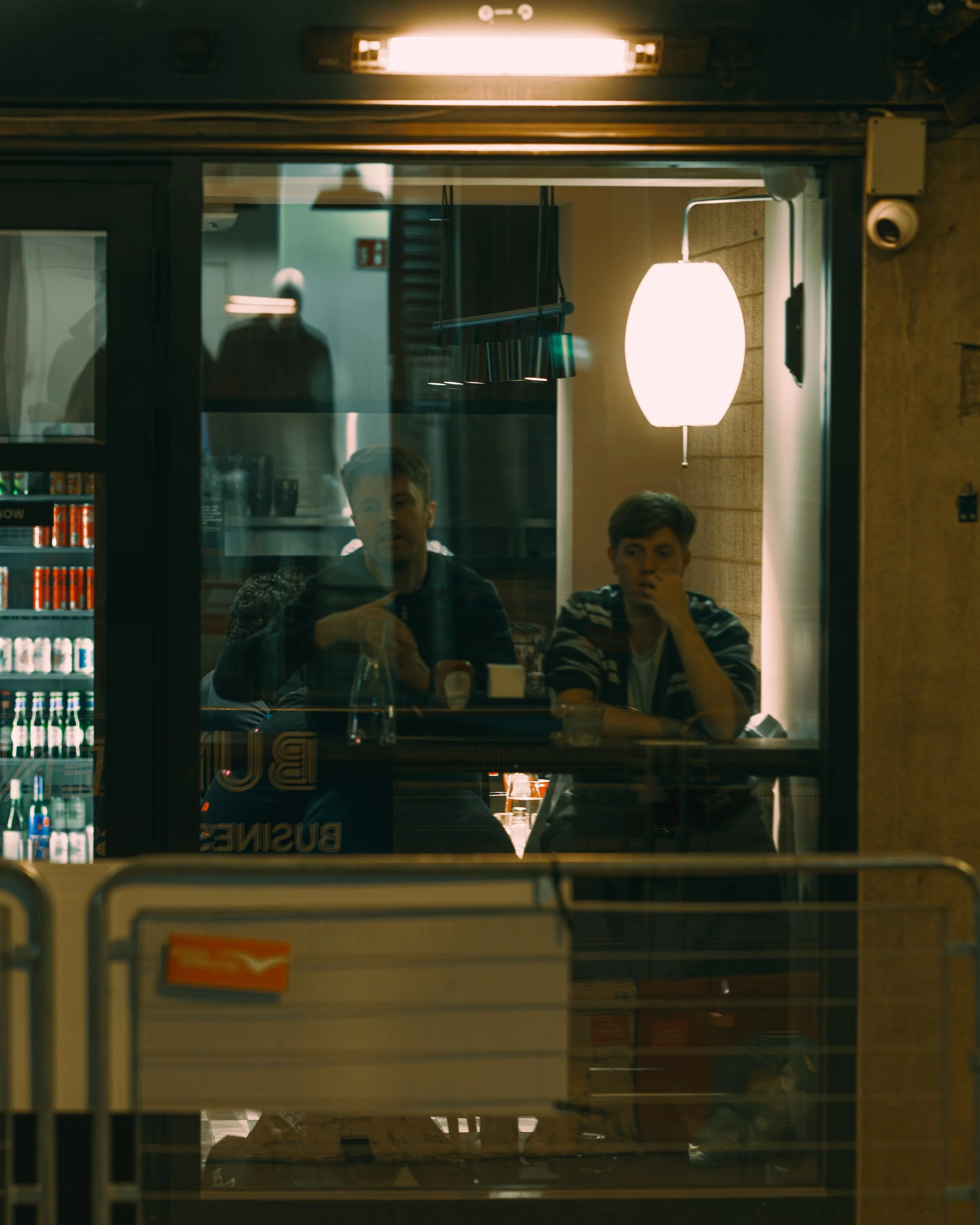 Two young men sitting inside a restaurant or cafe, seen through a large window at night. One is wearing a striped shirt, with his hand on his chin; the other has short hair and is looking forward.