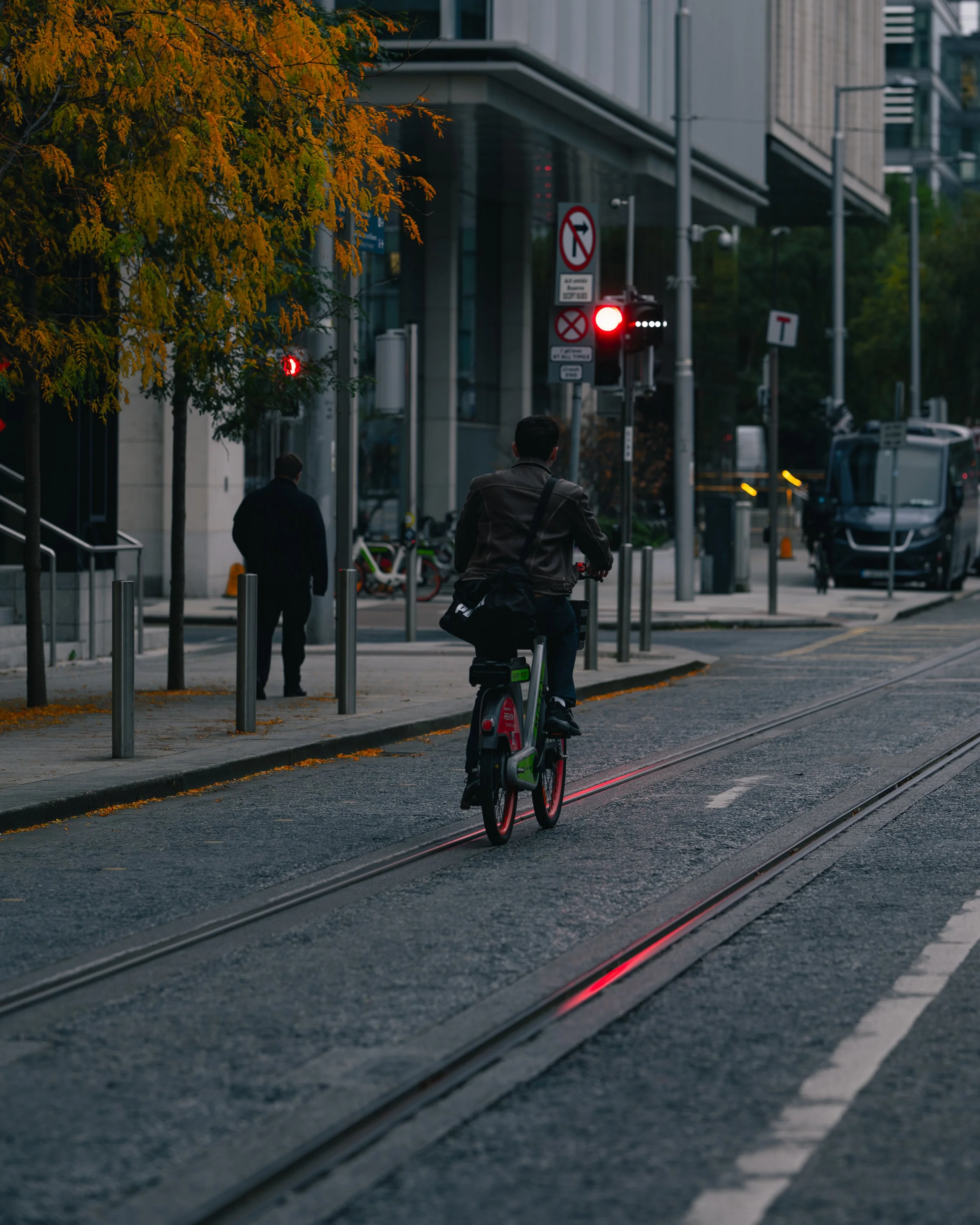 A person riding an electric bike on a city street with tram tracks, on a red light, with buildings and trees visible in the background.
