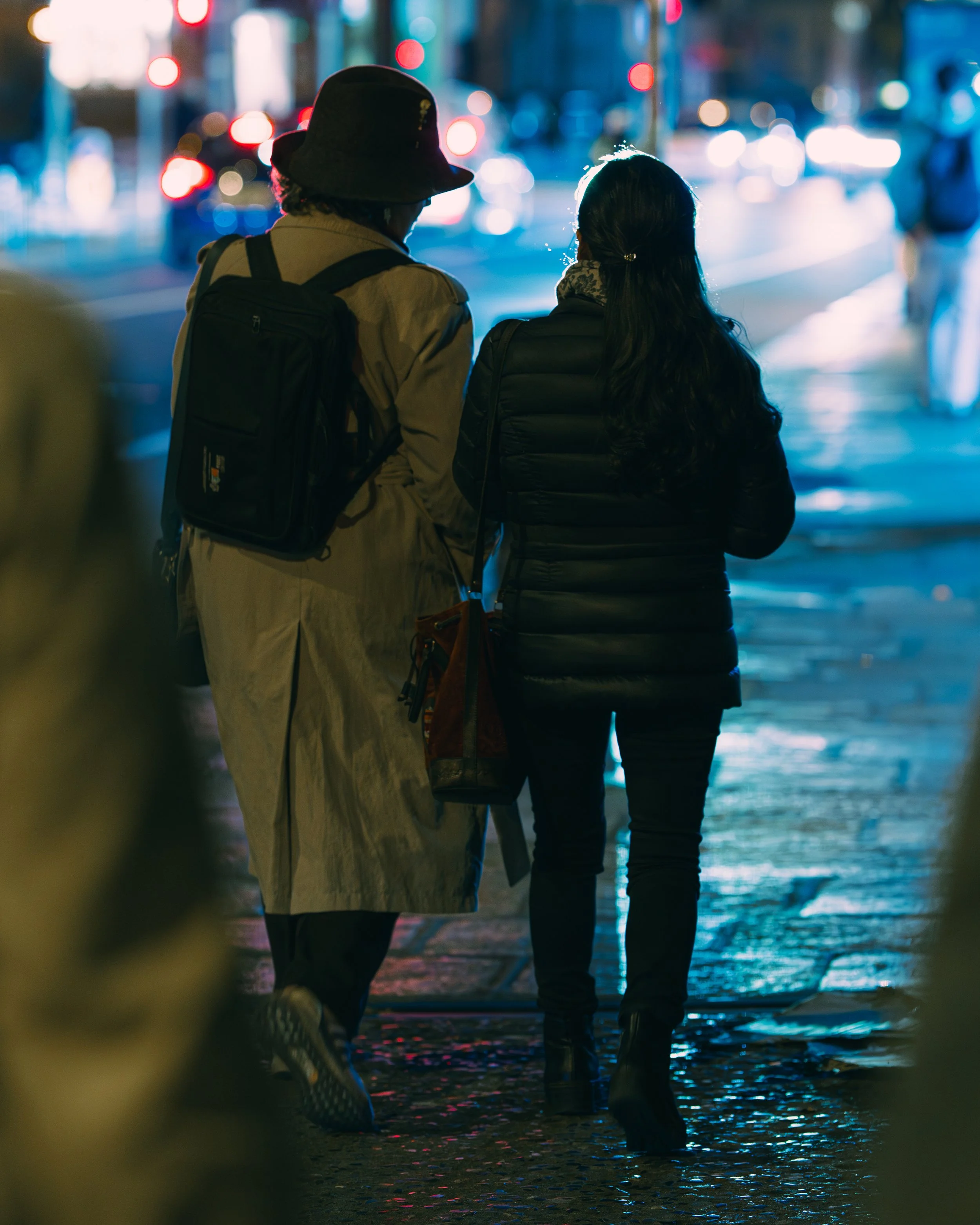 Two women with backpacks walking on a city sidewalk at night, illuminated by streetlights and traffic.
