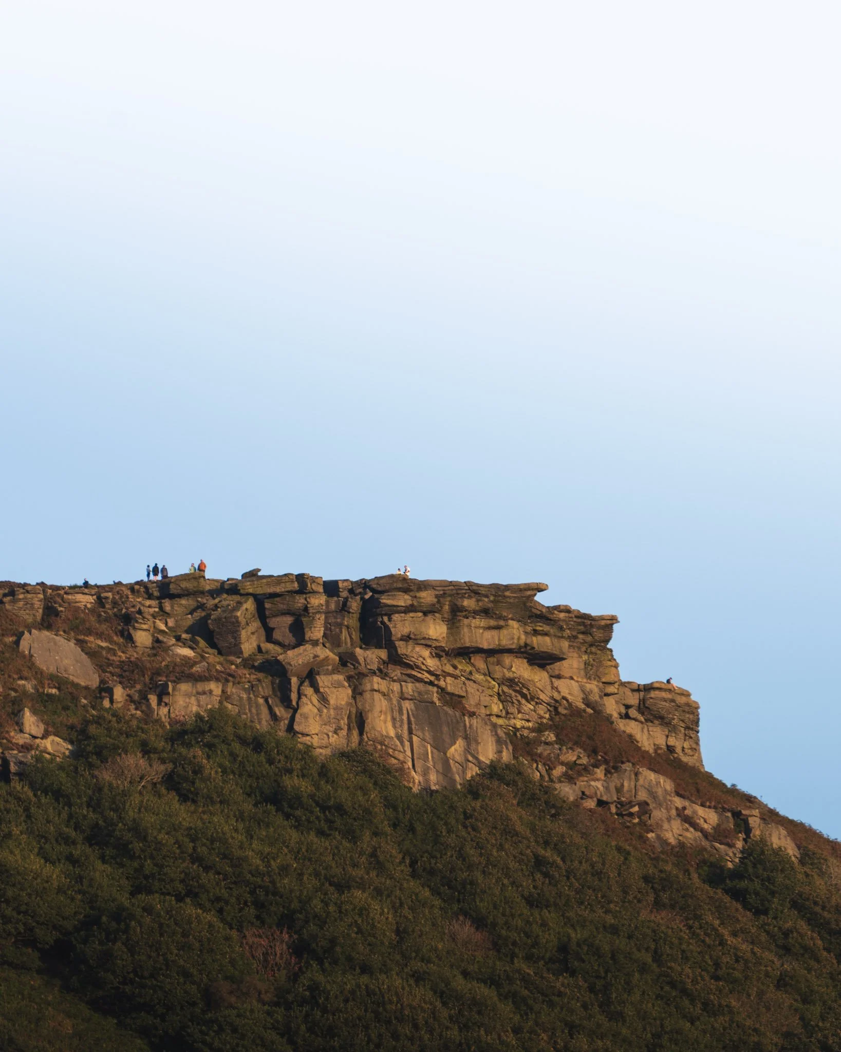 A rocky hill with a few people standing on top, surrounded by green vegetation, against a clear blue sky.