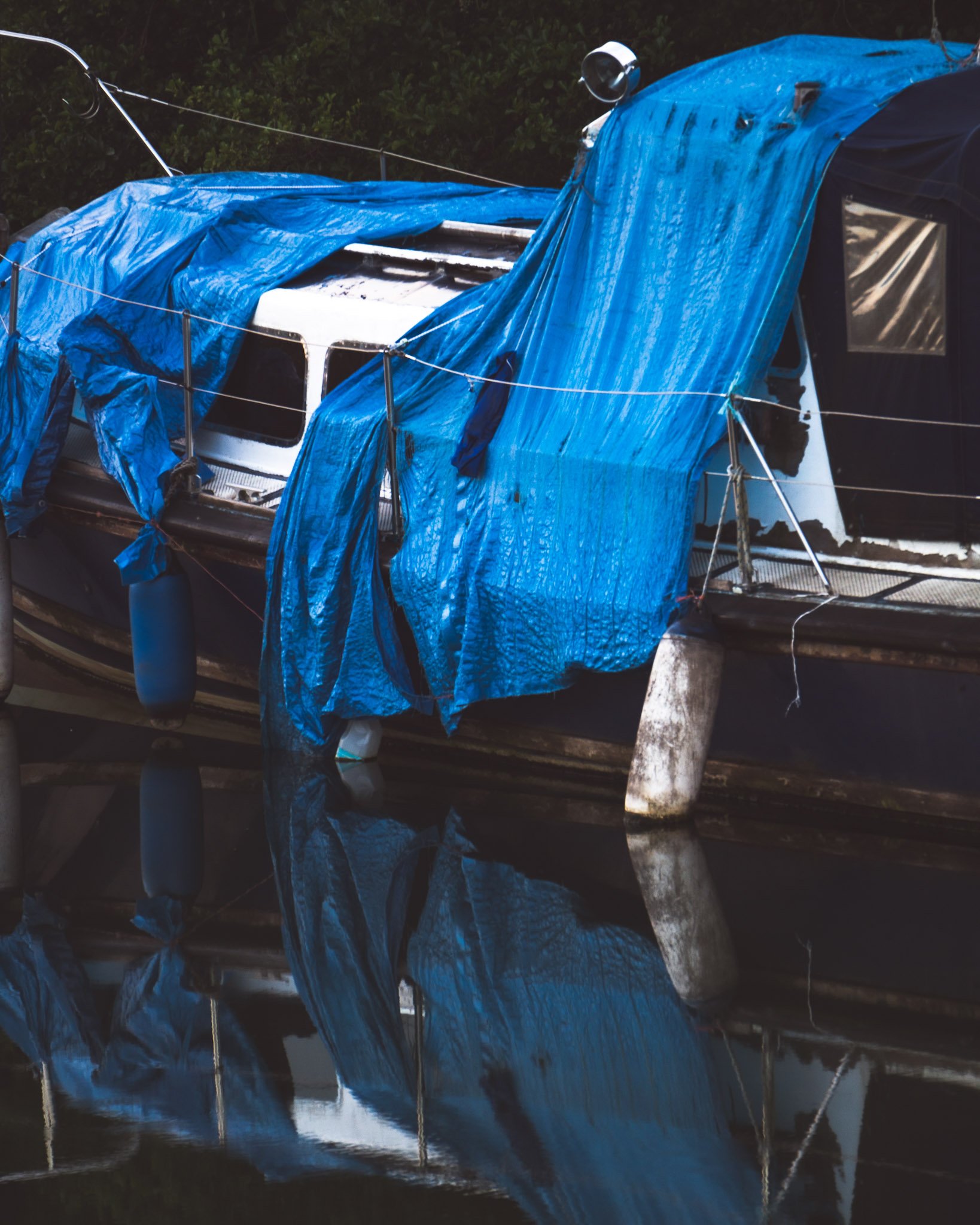 A boat covered with a blue tarp, moored at a dock, with its reflection visible in the water.