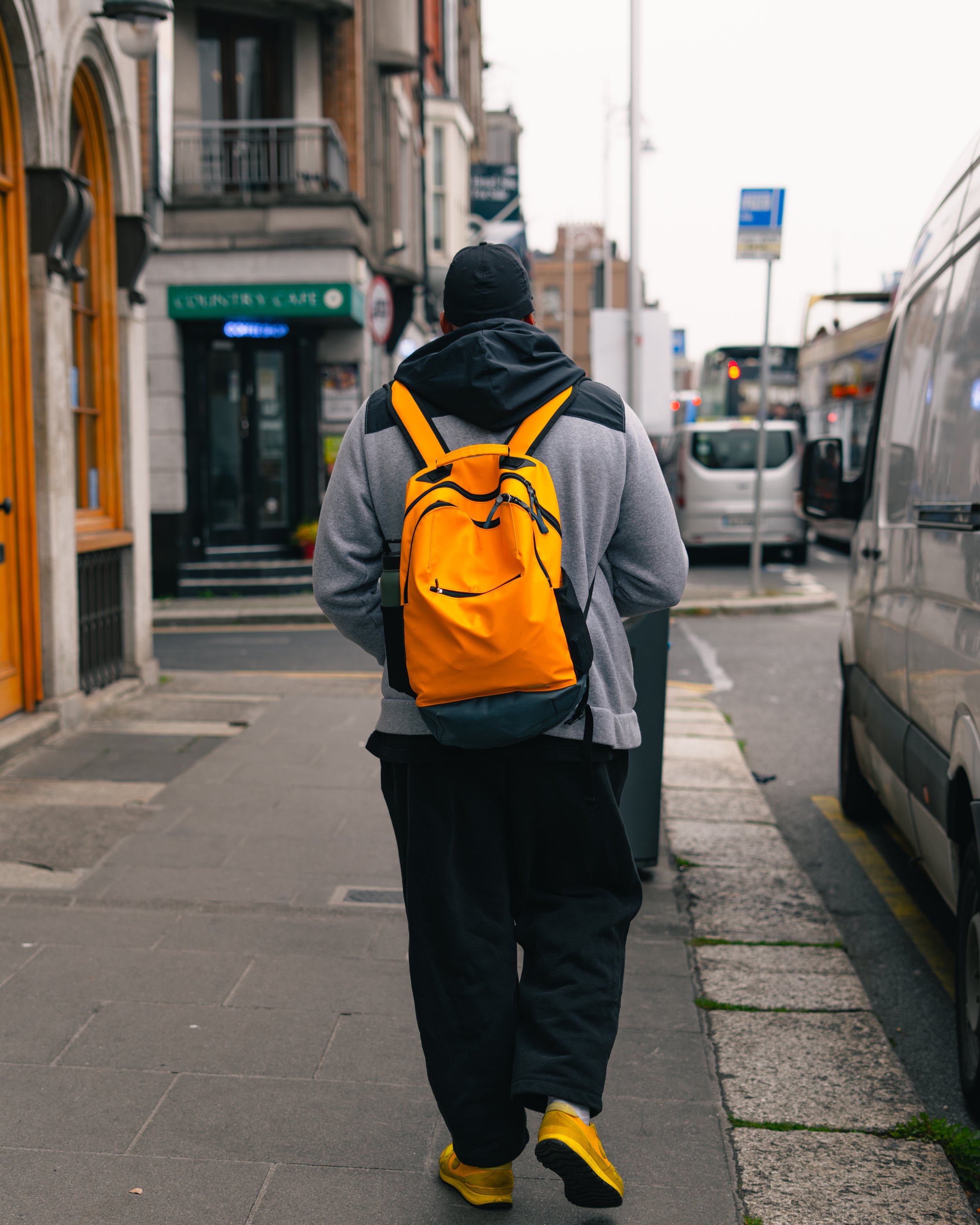 Person walking on city sidewalk with yellow backpack, gray and black hoodie, and yellow shoes.