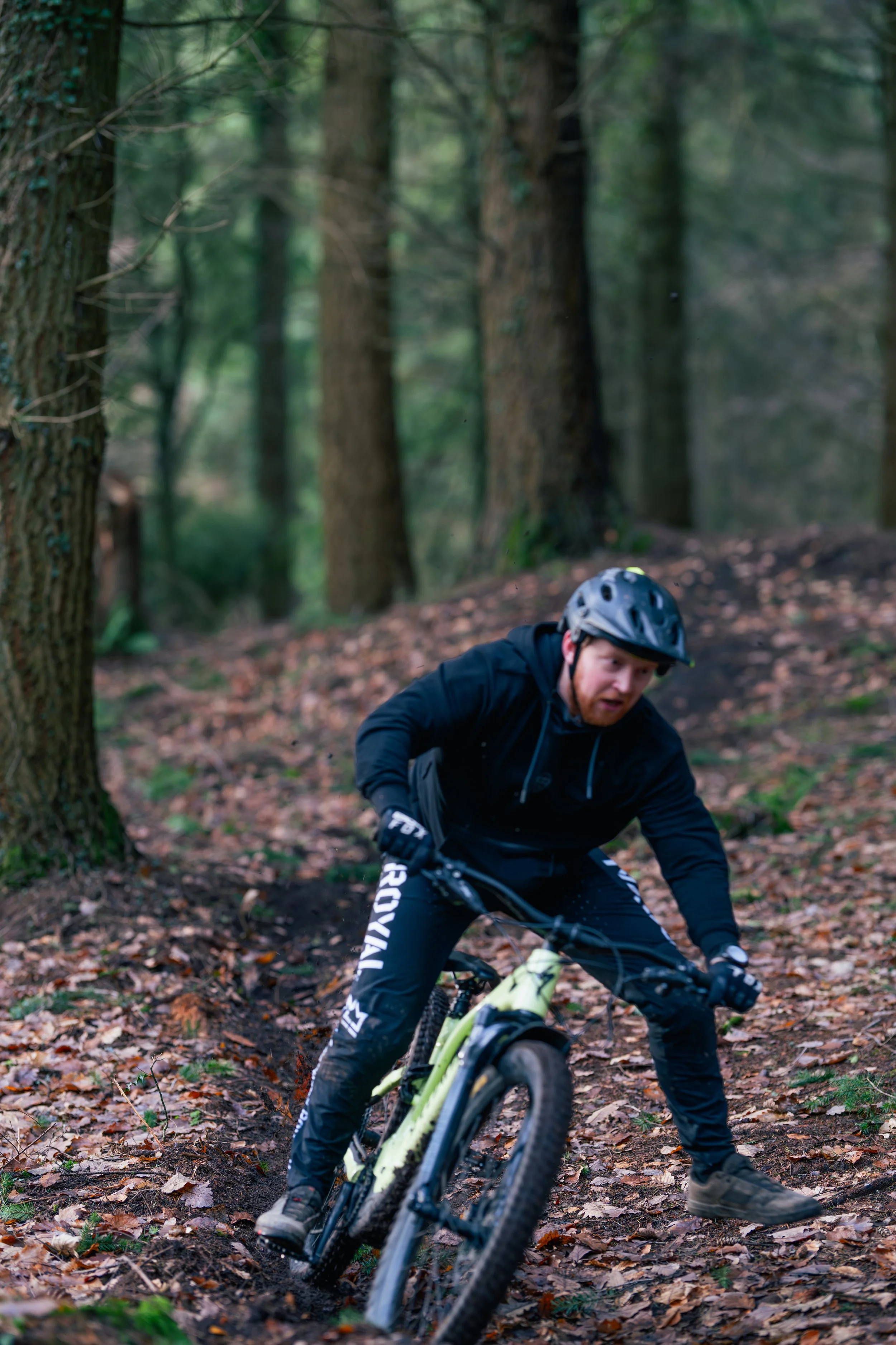 A man wearing a helmet and dark outdoor clothing riding a mountain bike on a forest trail covered with leaves.
