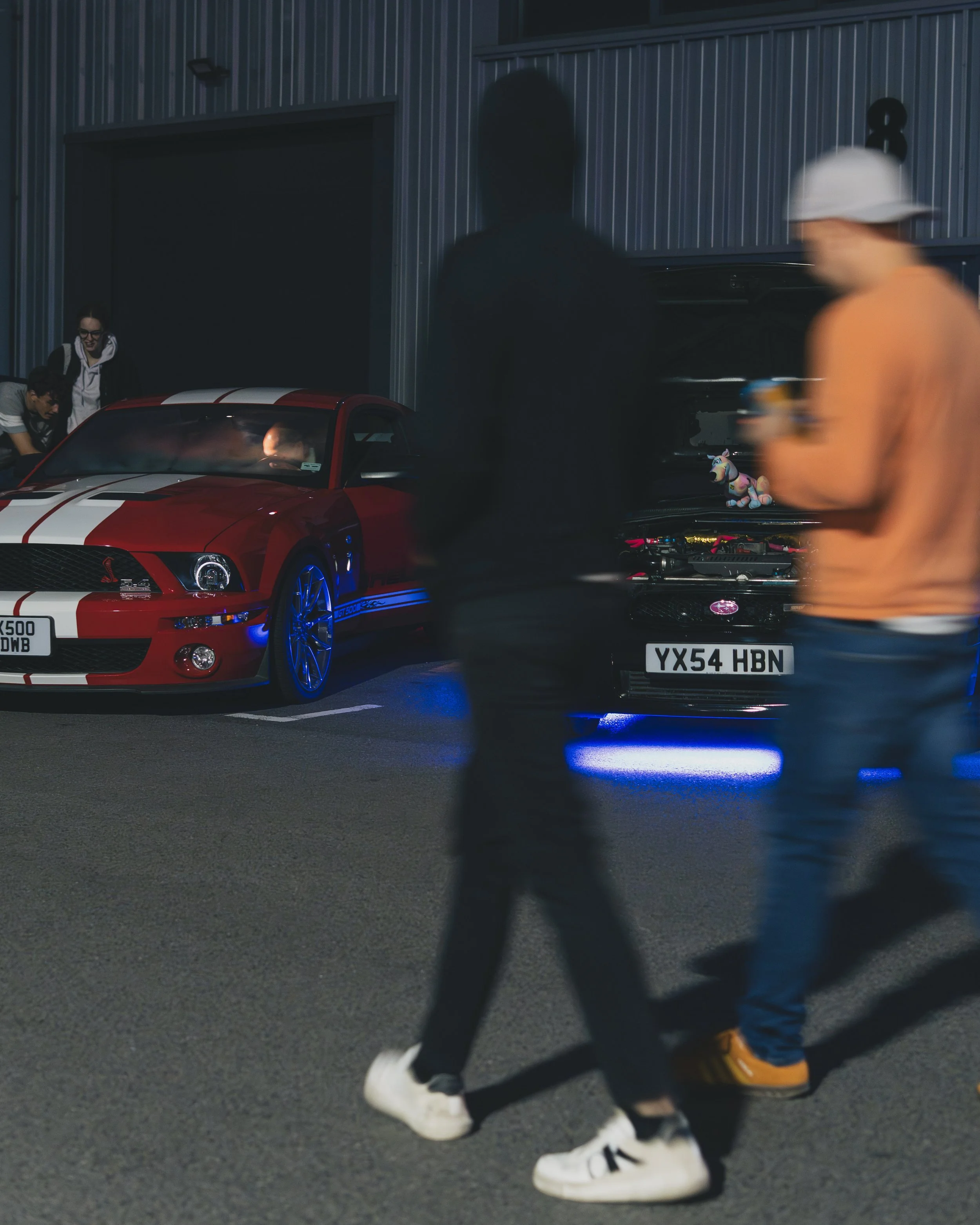 People walking past two cars, a red Ford Mustang Shelby GT500 with white racing stripes and an open hood showcasing a stuffed unicorn toy on the engine, parked outside a building with a metal exterior at night.
