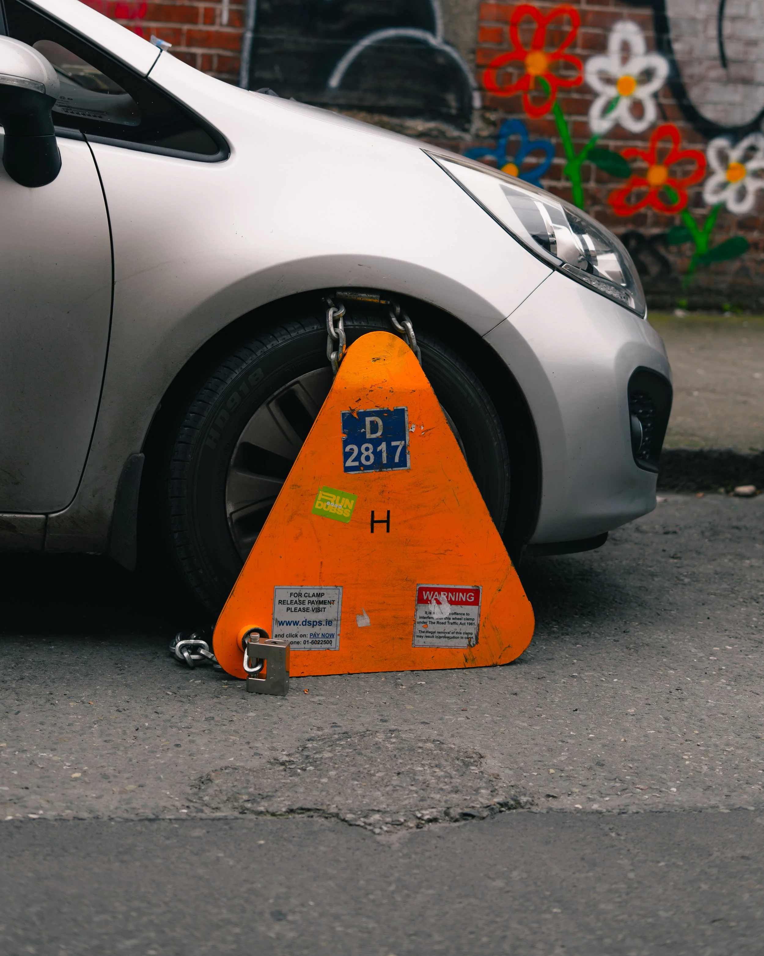 A car parked on the street with a bright orange wheel clamp attached to its front tire, preventing it from moving.