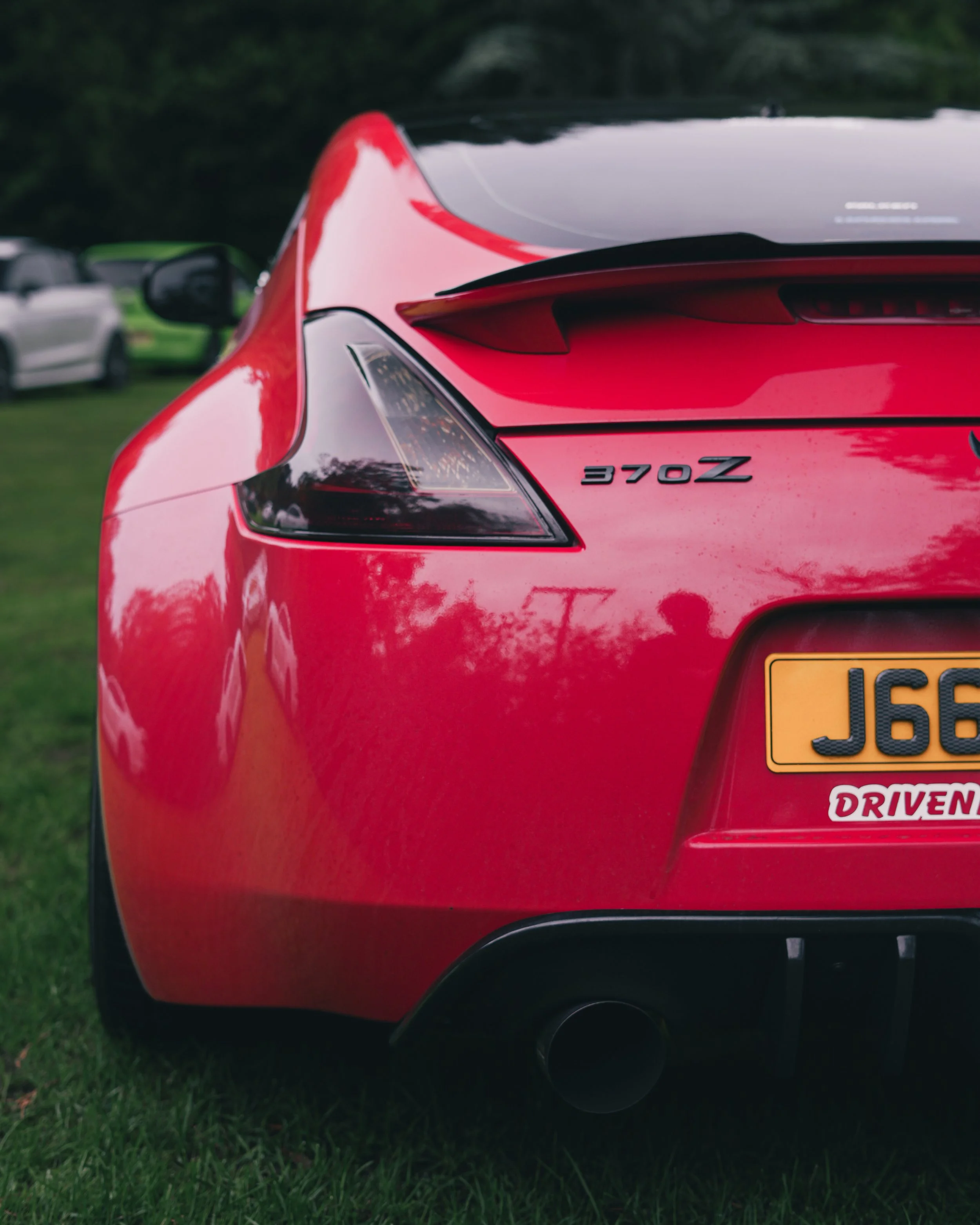 Close-up of the rear of a red  Nissan z370 , showing the taillight, badge that reads '370Z', and a yellow license plate with a sticker that says 'DRIVEN'.