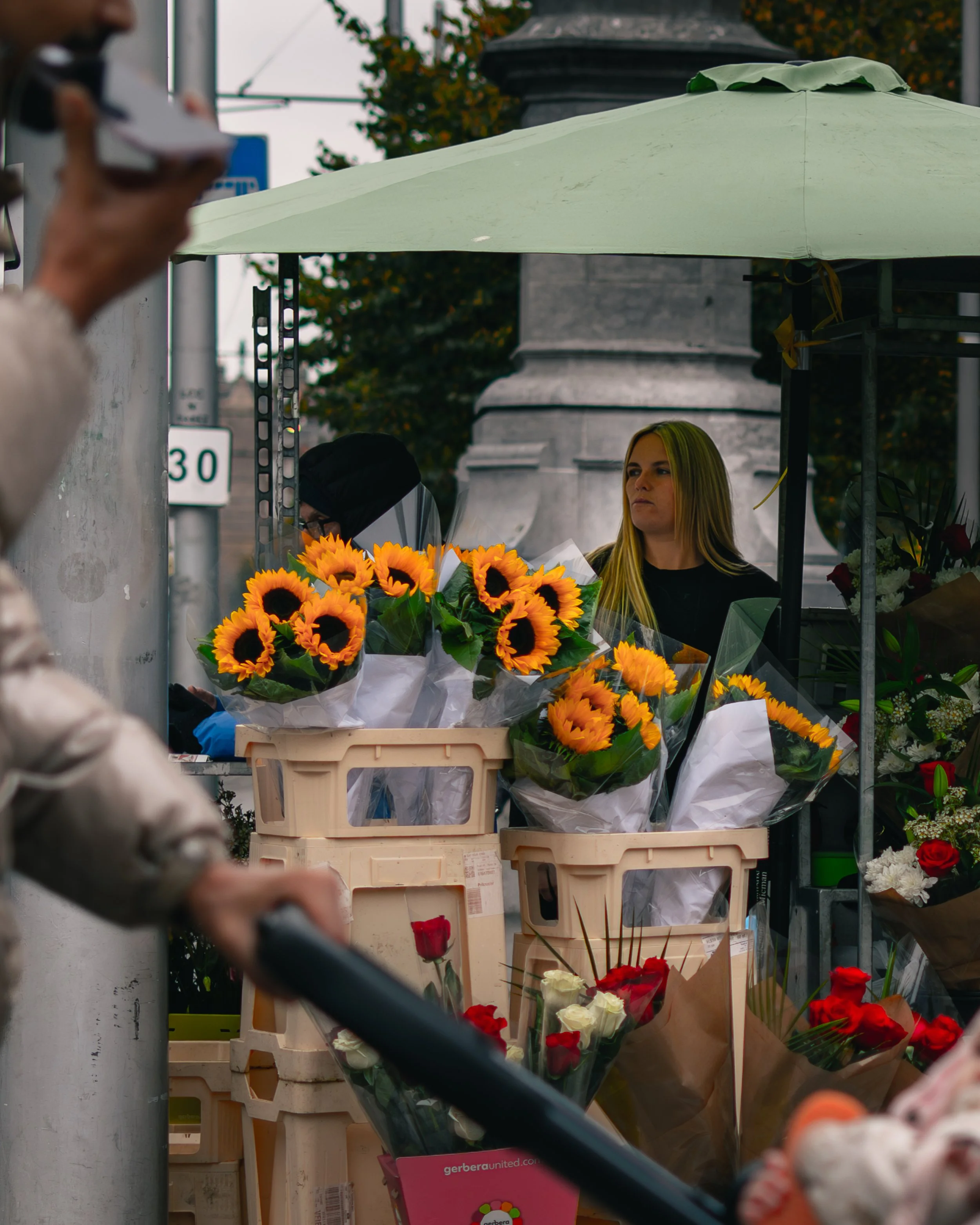 A flower vendor stand with bouquets of sunflowers and roses, a woman with long blonde hair looking to the side, and a person in the foreground pushing a shopping cart, outdoors under a green umbrella